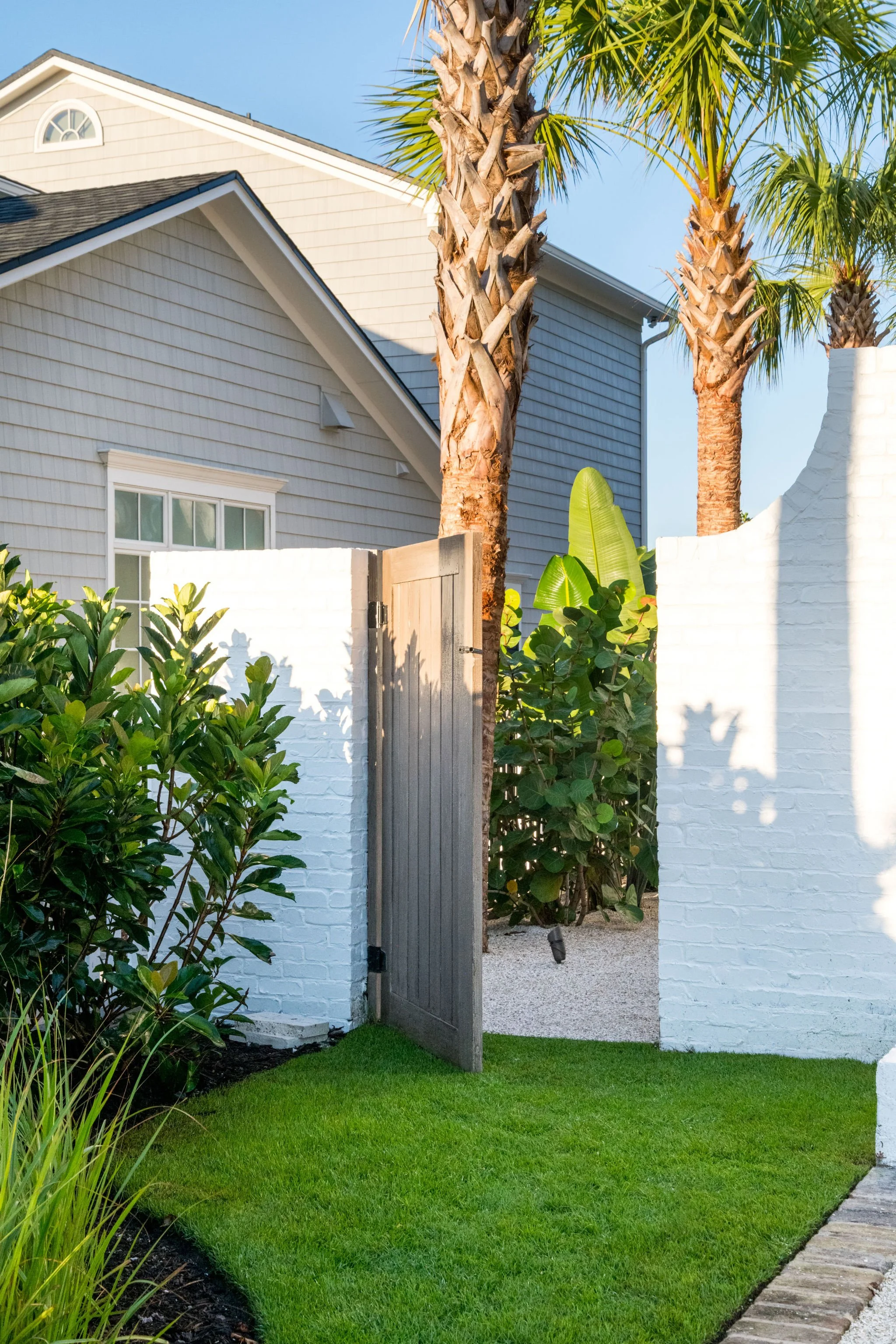 Custom wood garden gate framed by curved white brick walls, tropical palms, and lush evergreen plantings.