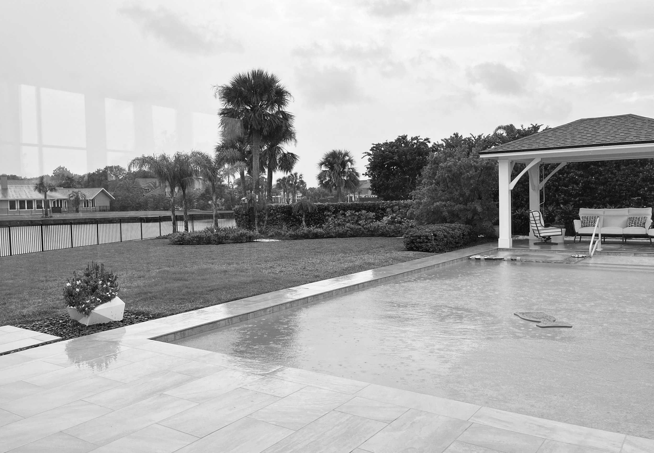 A backyard pool area with a covered seating area, palm trees, a lawn, and a fence surrounding a body of water, prompting a cloudy sky.