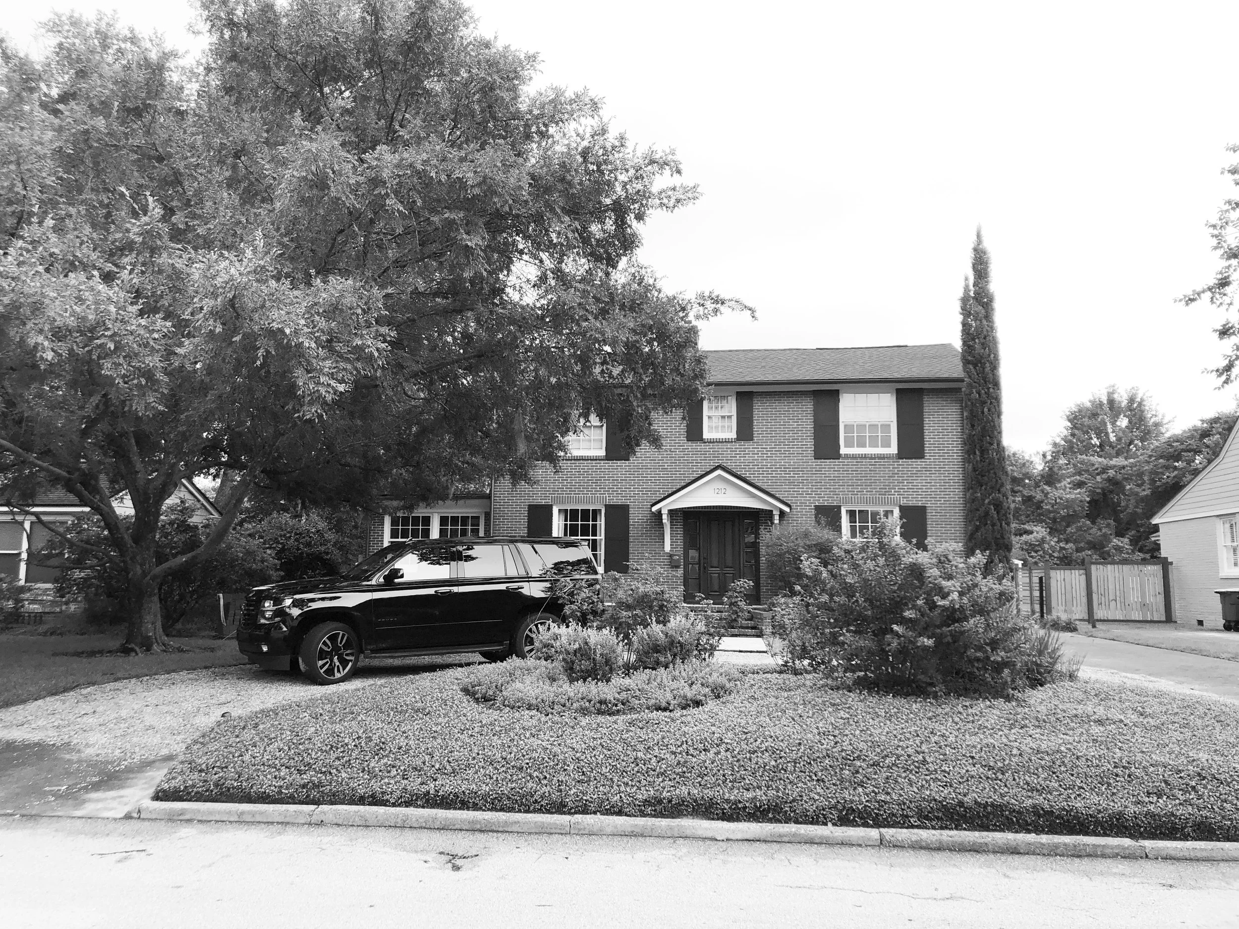 A black SUV parked in front of a two-story brick house with a front yard, large trees, and shrubs, during daytime.