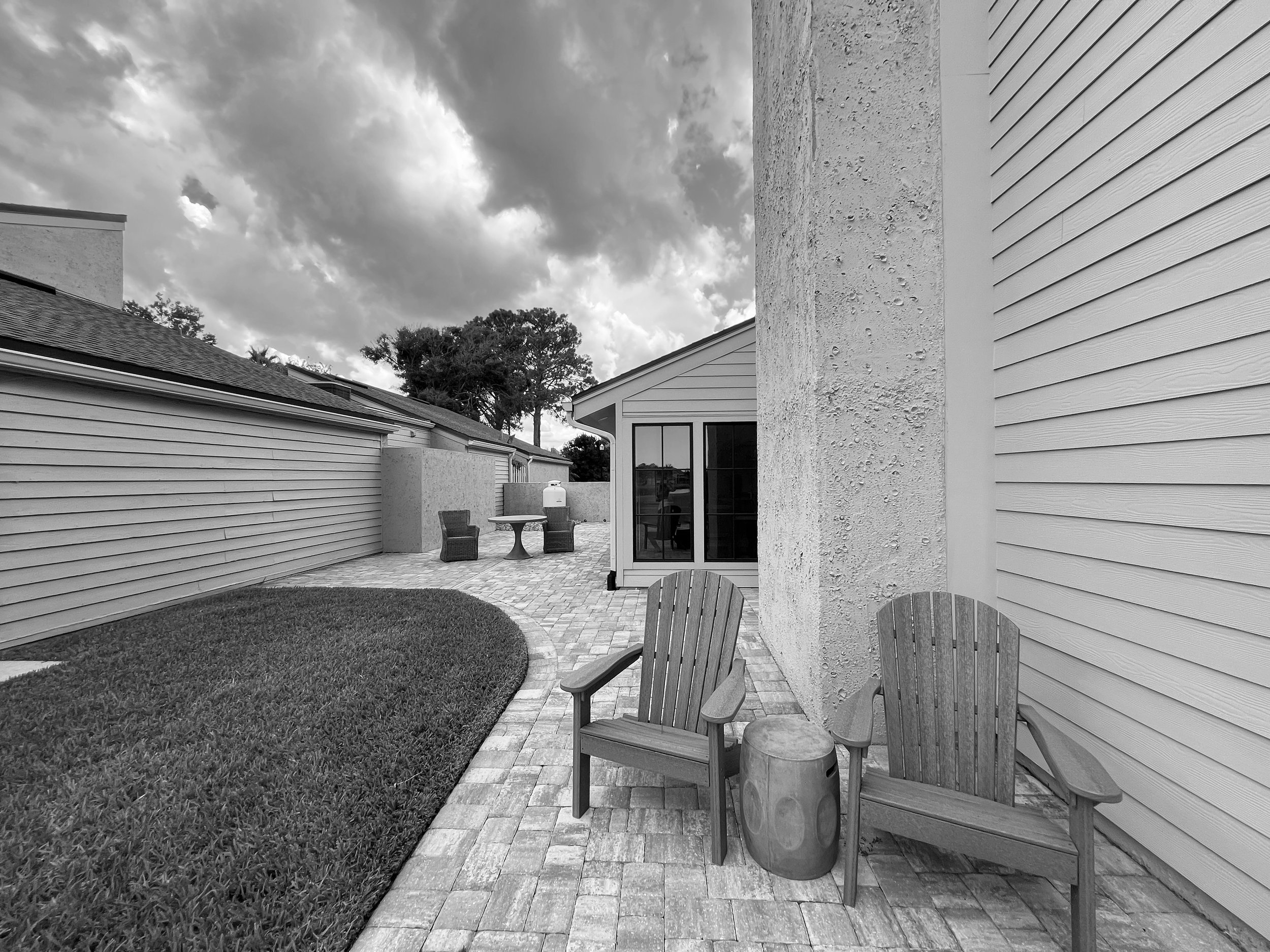 Backyard patio with wooden chairs, a small table, and a brick pathway, with a lawn and a cloudy sky.