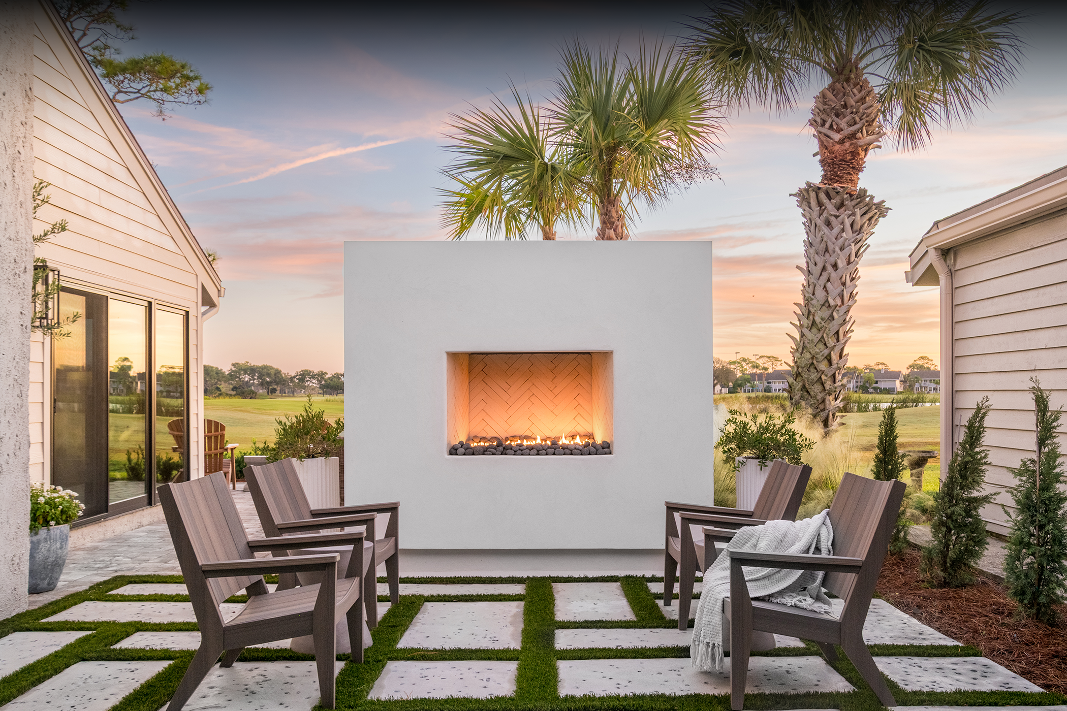 Outdoor patio with wooden chairs around a modern square fireplace, palm trees, potted plants, and a sunset sky.