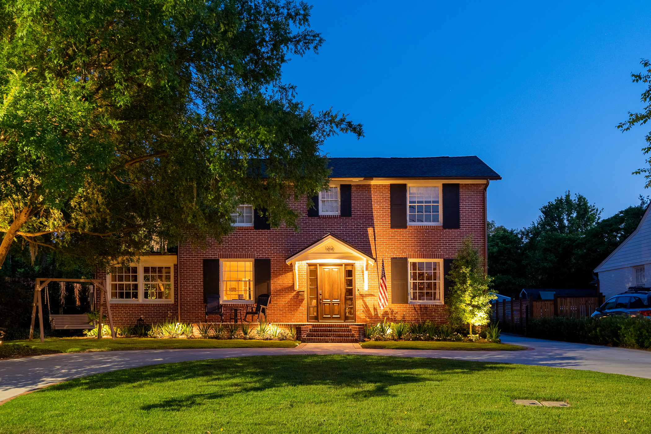 A two-story brick house with a front porch lit at dusk, surrounded by a green lawn and trees, with a swing set on the left and a driveway on the right.