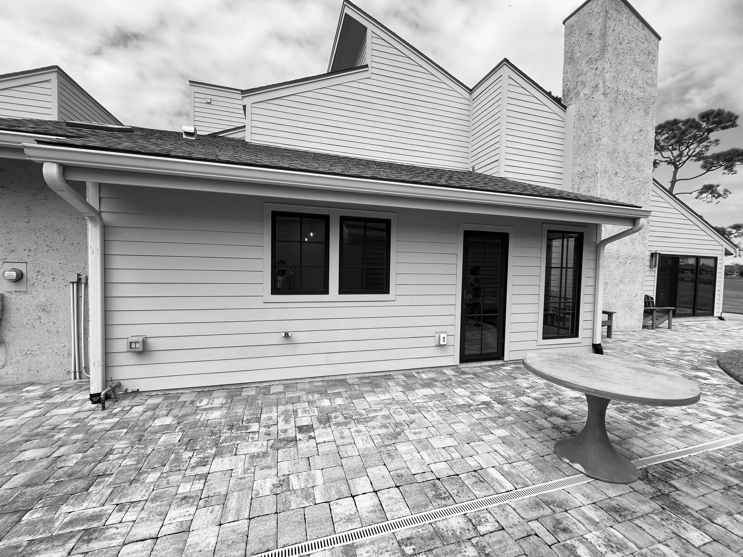 Backyard patio with brick pavers, a round table, and a house with siding, windows, sliding door, chimney, and neighboring house. Overcast sky.