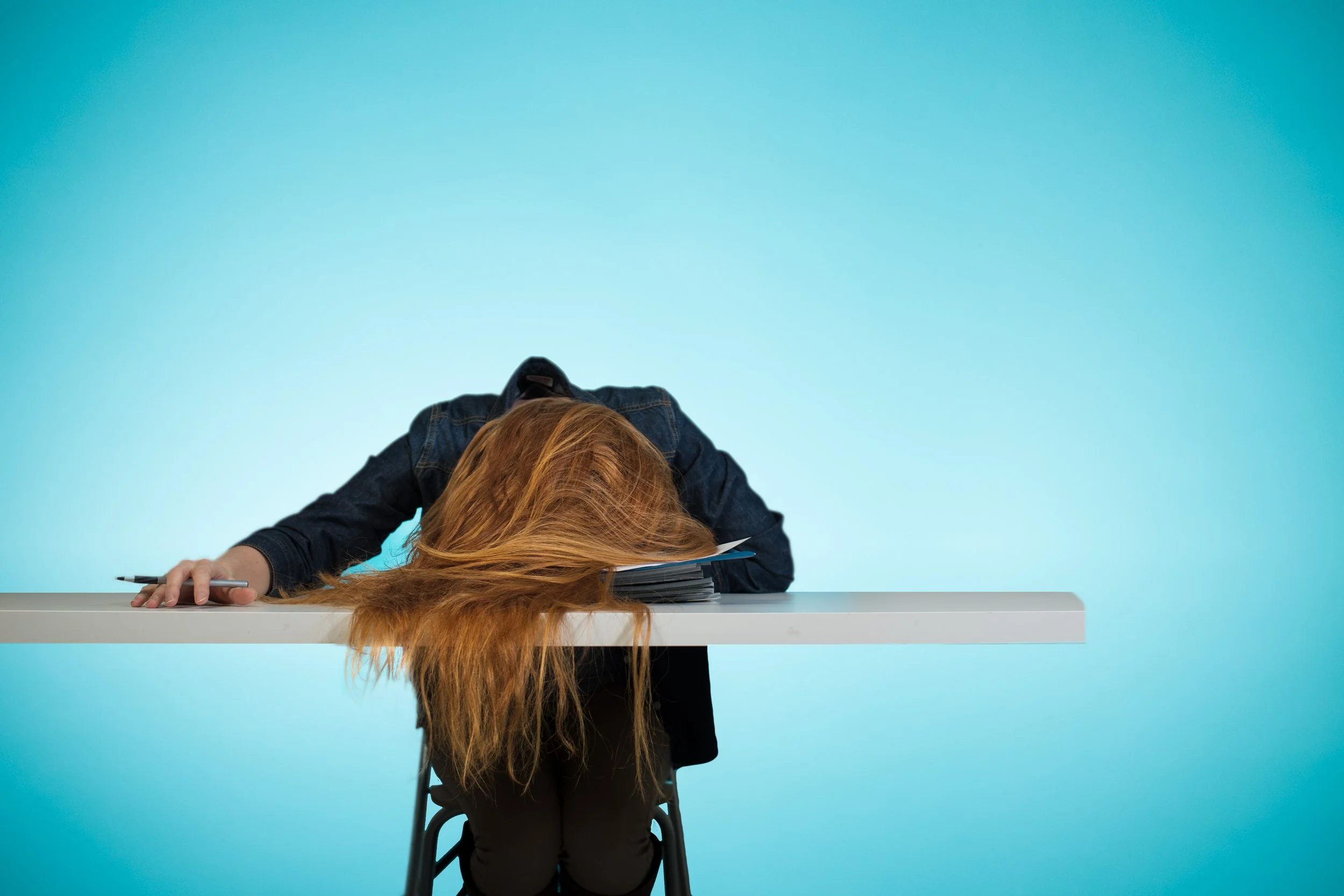 Lauren Westfall slumped over a table with her head on the surface with a pen in her hand and papers in front of her, against a blue background.