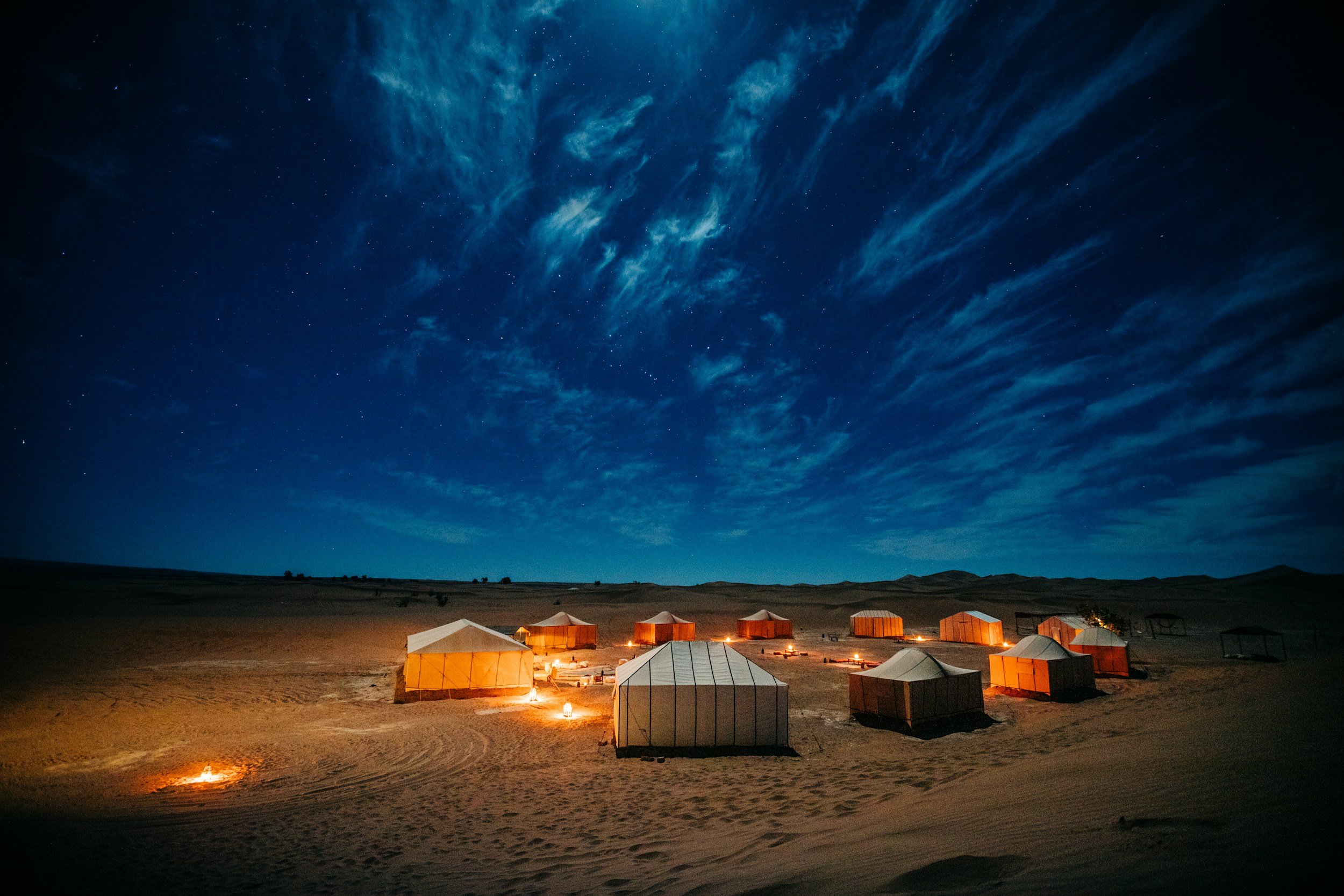 Desert campsite at night with multiple tents illuminated by small fires, under a starry sky with wispy clouds.