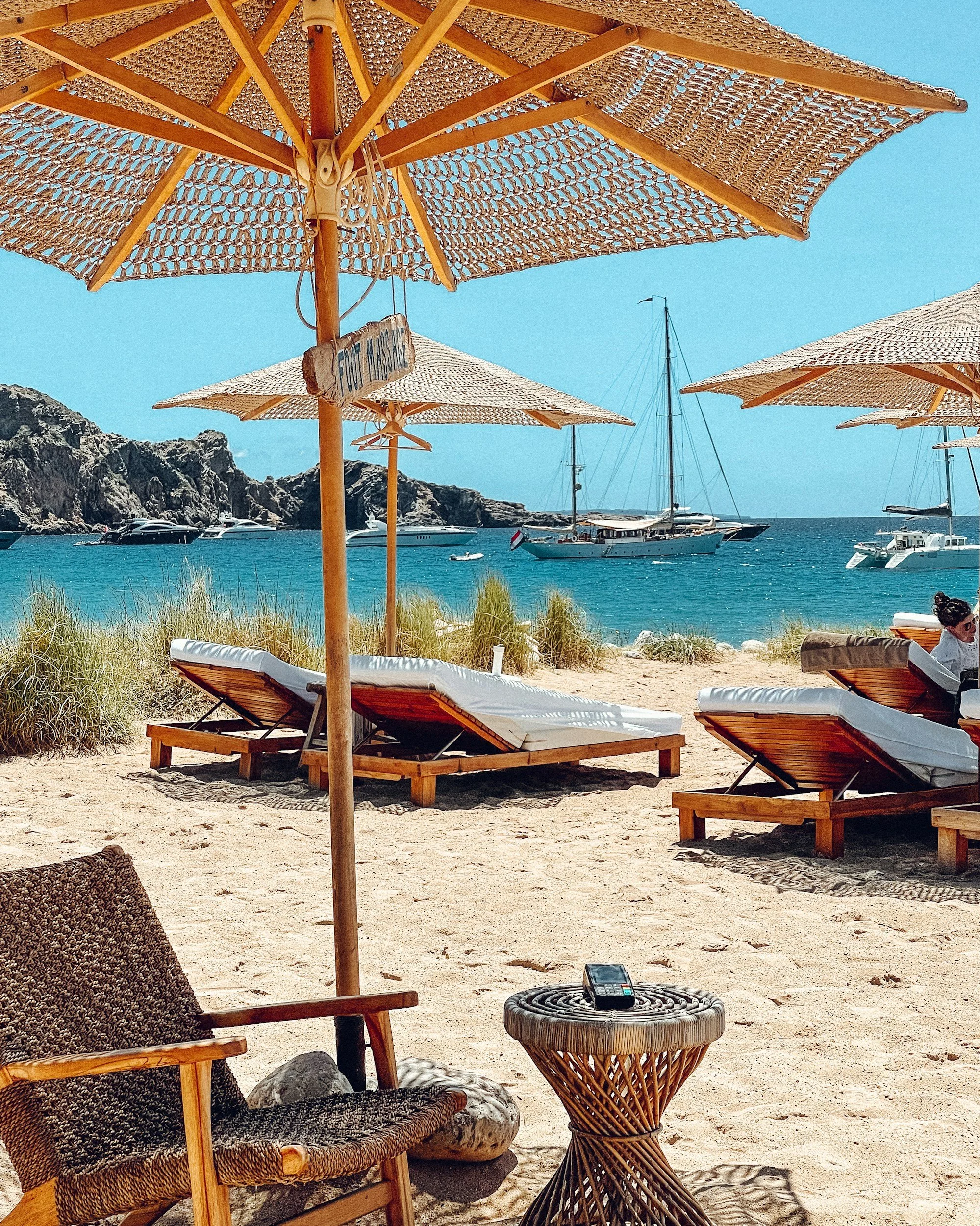 A beach scene with wooden lounge chairs and large woven umbrellas on sandy shore, sailboats and yachts anchored in clear blue water, rocky cliffs in background, and a woman relaxing on a lounge chair.