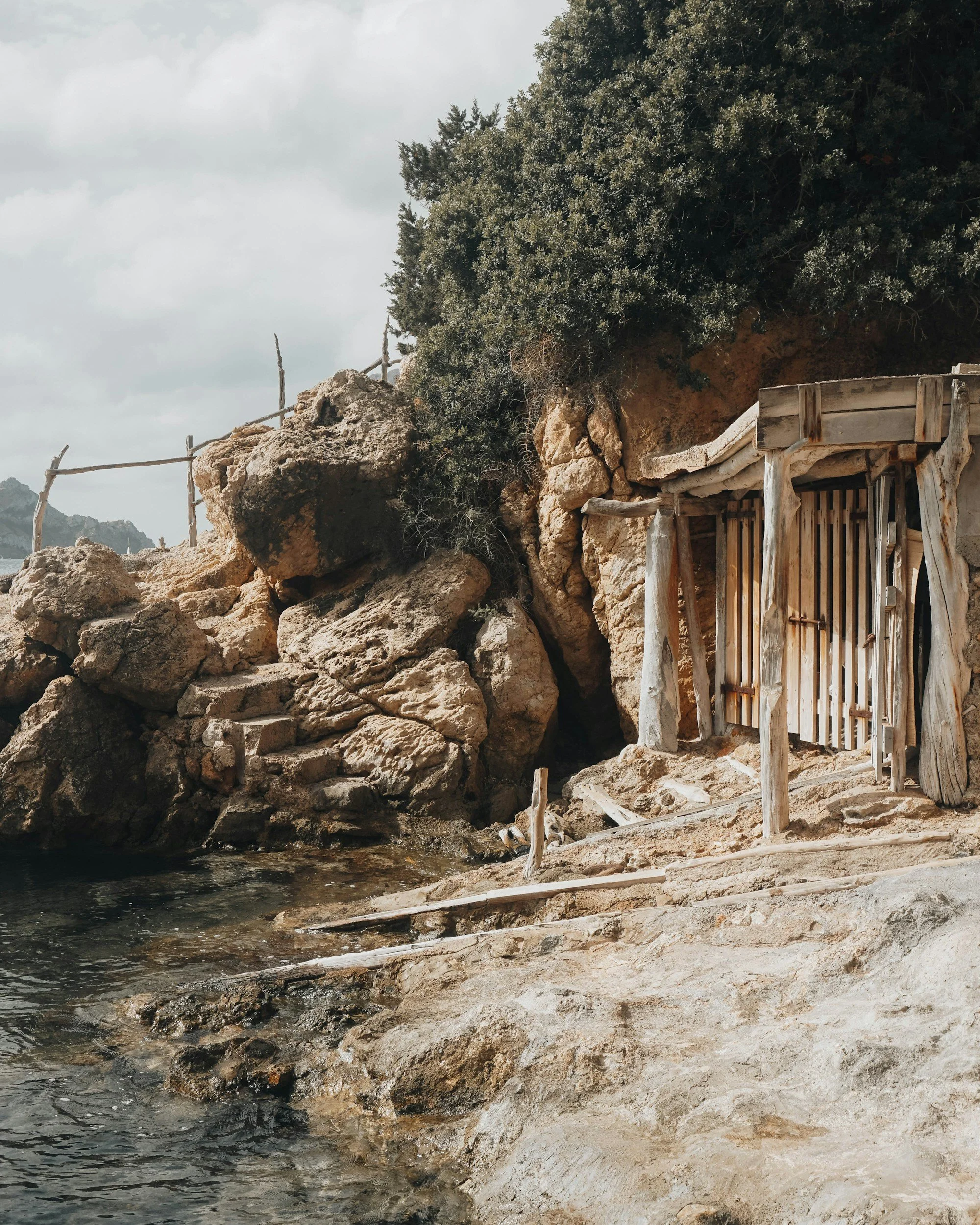 A rustic wooden door built into a rocky hillside near water, with large rocks and greenery nearby.