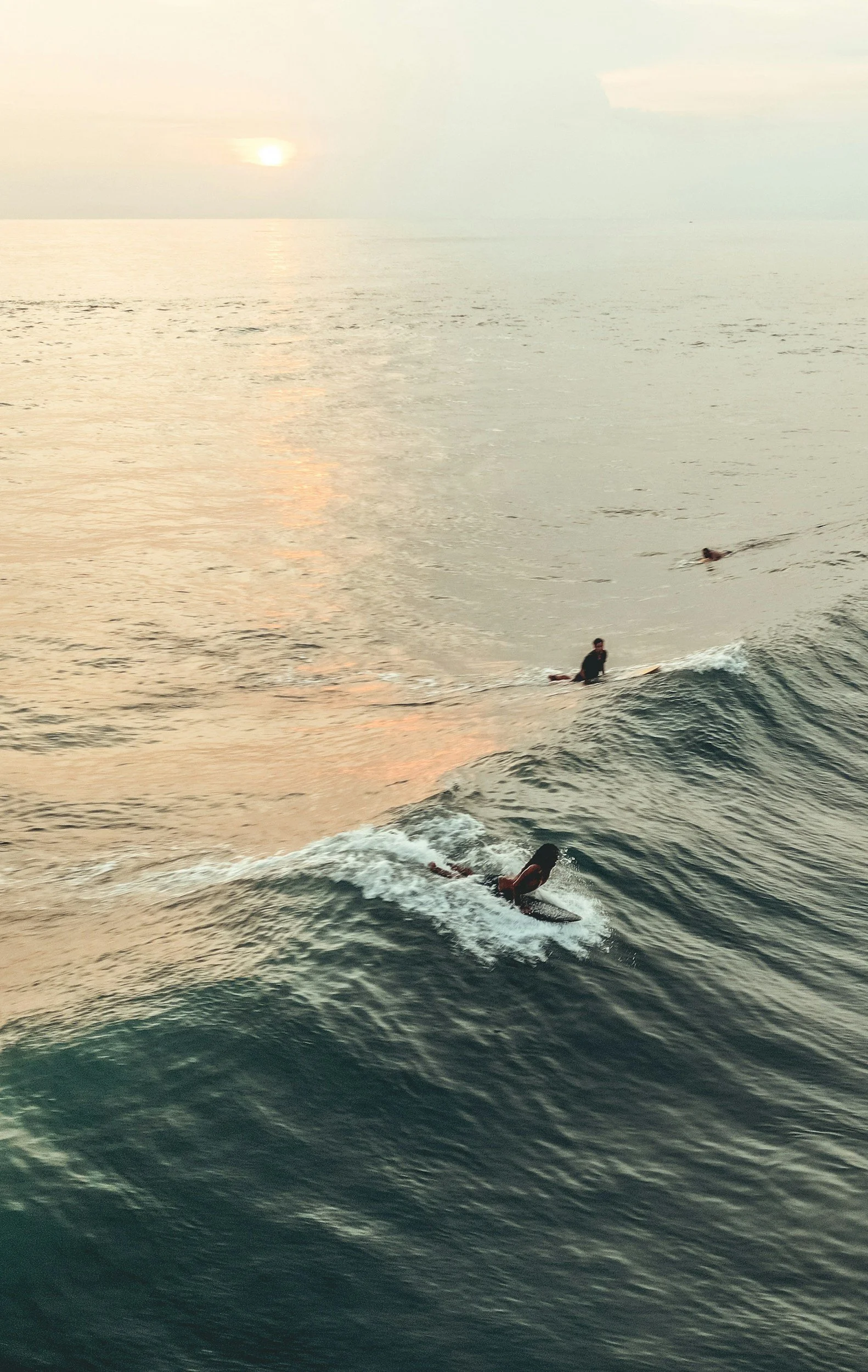 Three surfers riding waves on the ocean at sunset, with the sun setting on the horizon.