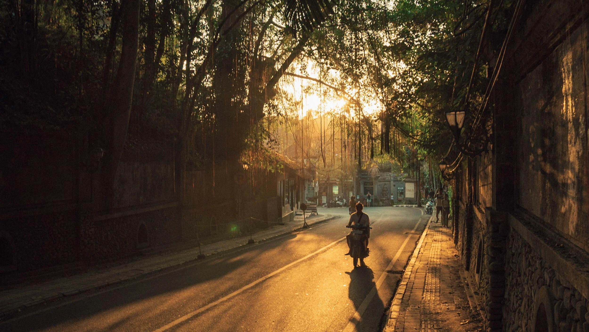 A street scene at sunset with people walking and riding motorbikes. The street is bordered by a stone wall on the right and trees overhead, casting shadows. The sunlight filters through the foliage.