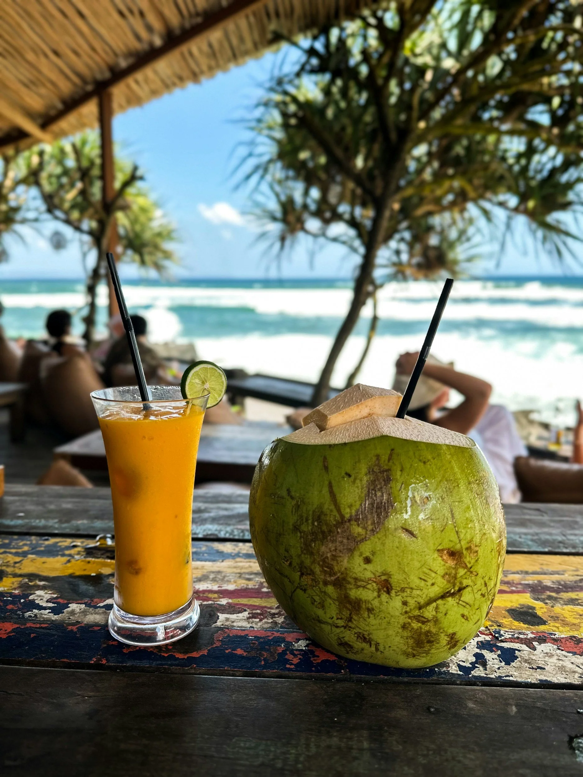 A tropical beachside bar with a colorful wooden table holding a glass of orange juice with a lime slice and a green coconut with a straw. In the background, there are people relaxing under a thatched roof with the ocean and waves visible.