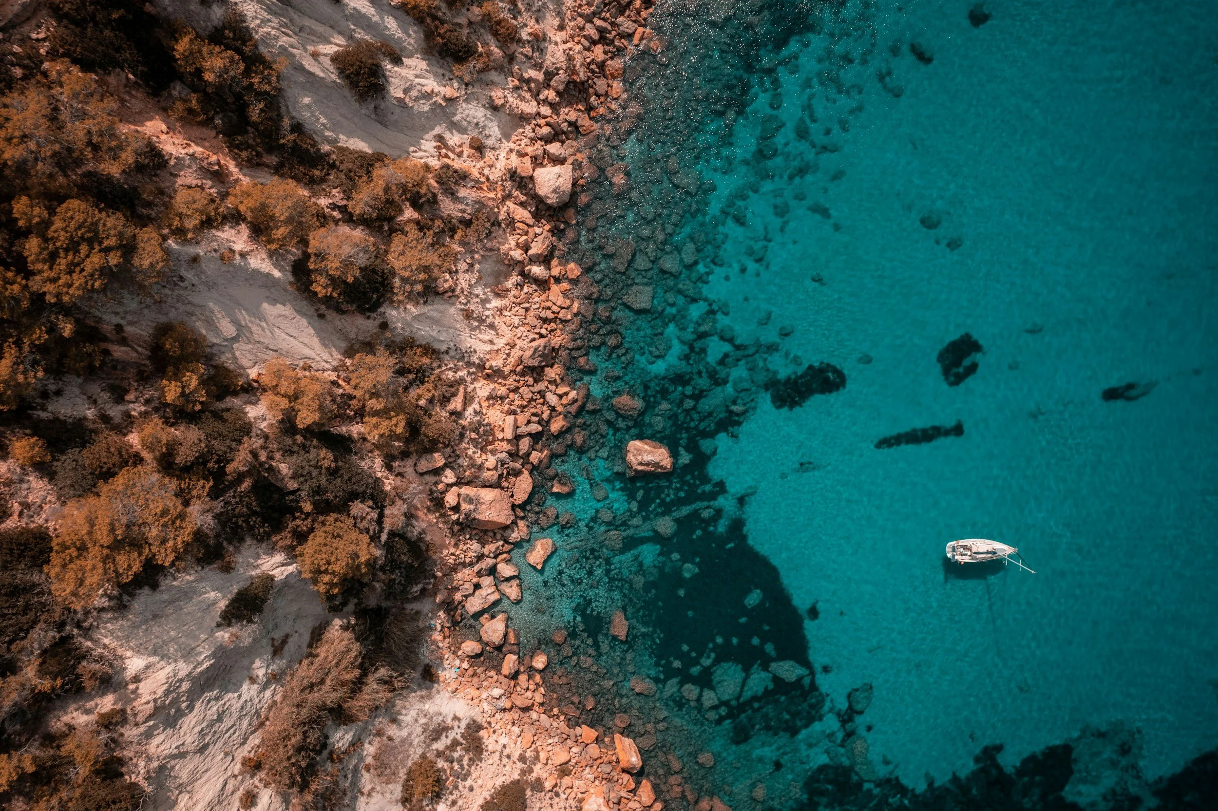 An aerial view of a rocky coastline with a boat anchored near the shore, surrounded by trees and clear blue water.
