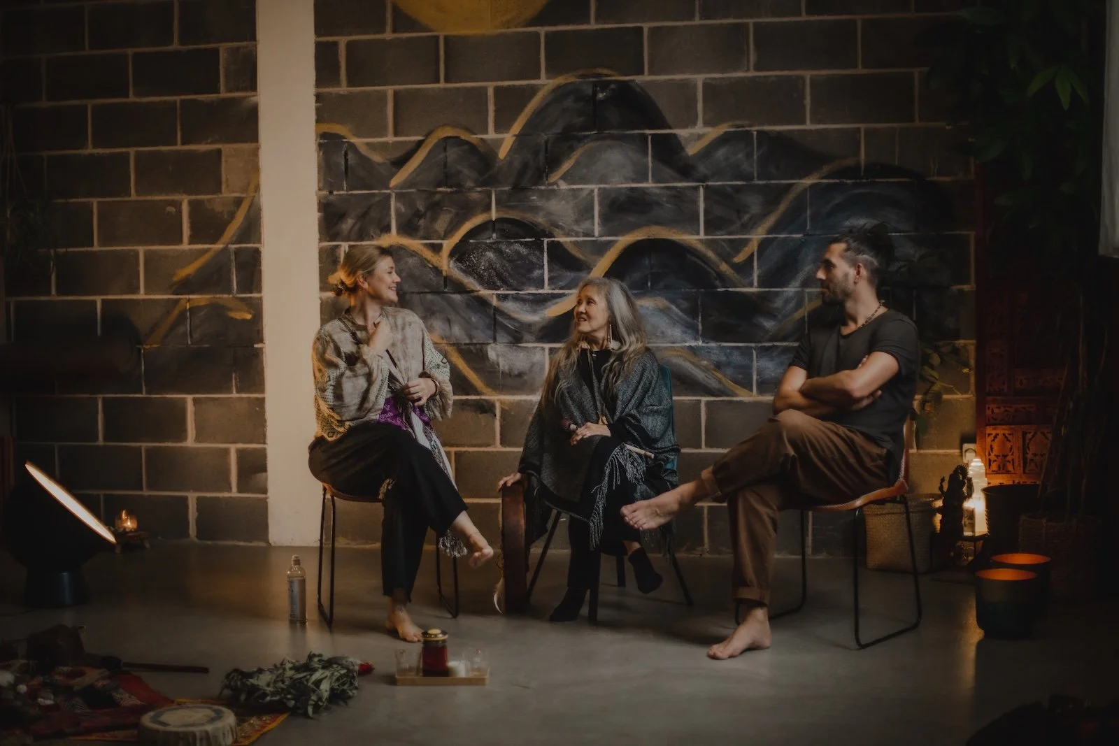 Three people having a conversation in a cozy, dimly-lit room with a black brick wall and mountain mural in the background. The woman on the left is smiling and talking, the middle woman is listening, and the man on the right is sitting with crossed arms. There are various items on the floor including candles, bottles, and cloths.