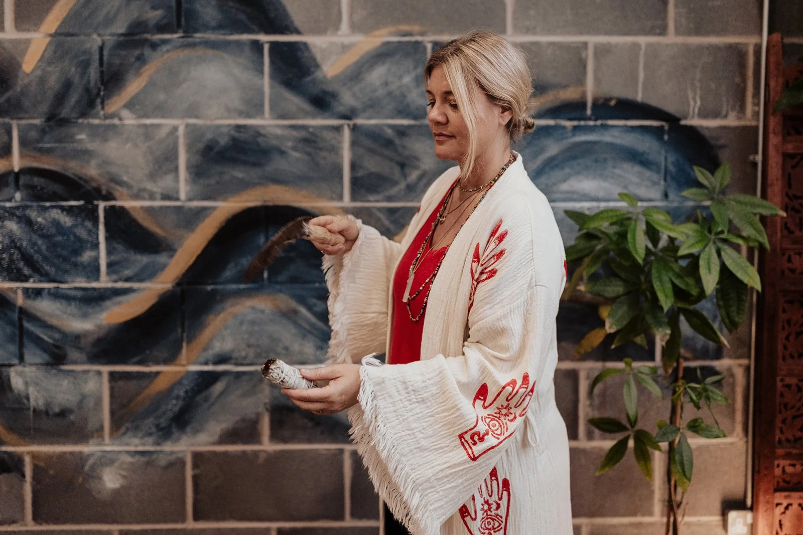 A woman in a white and red embroidered kimono holding a shell and a feather standing in front of a mural with blue, black, and beige swirls, next to a green potted plant.