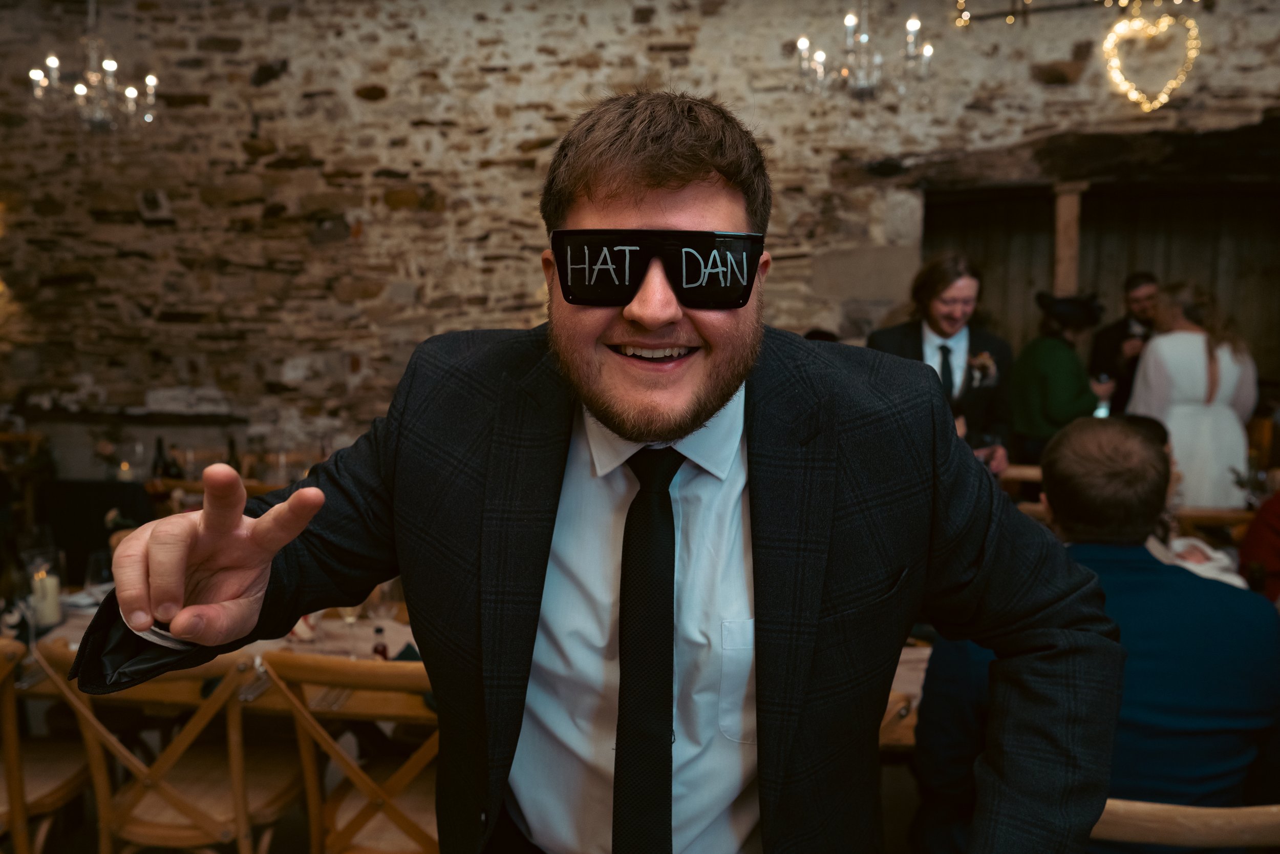 A man in a tuxedo smiling at a wedding celebration, wearing black glasses with the words 'HAT DAN' written on them in colorful marker, making a peace sign with his fingers, in a rustic indoor venue with guests in the background.