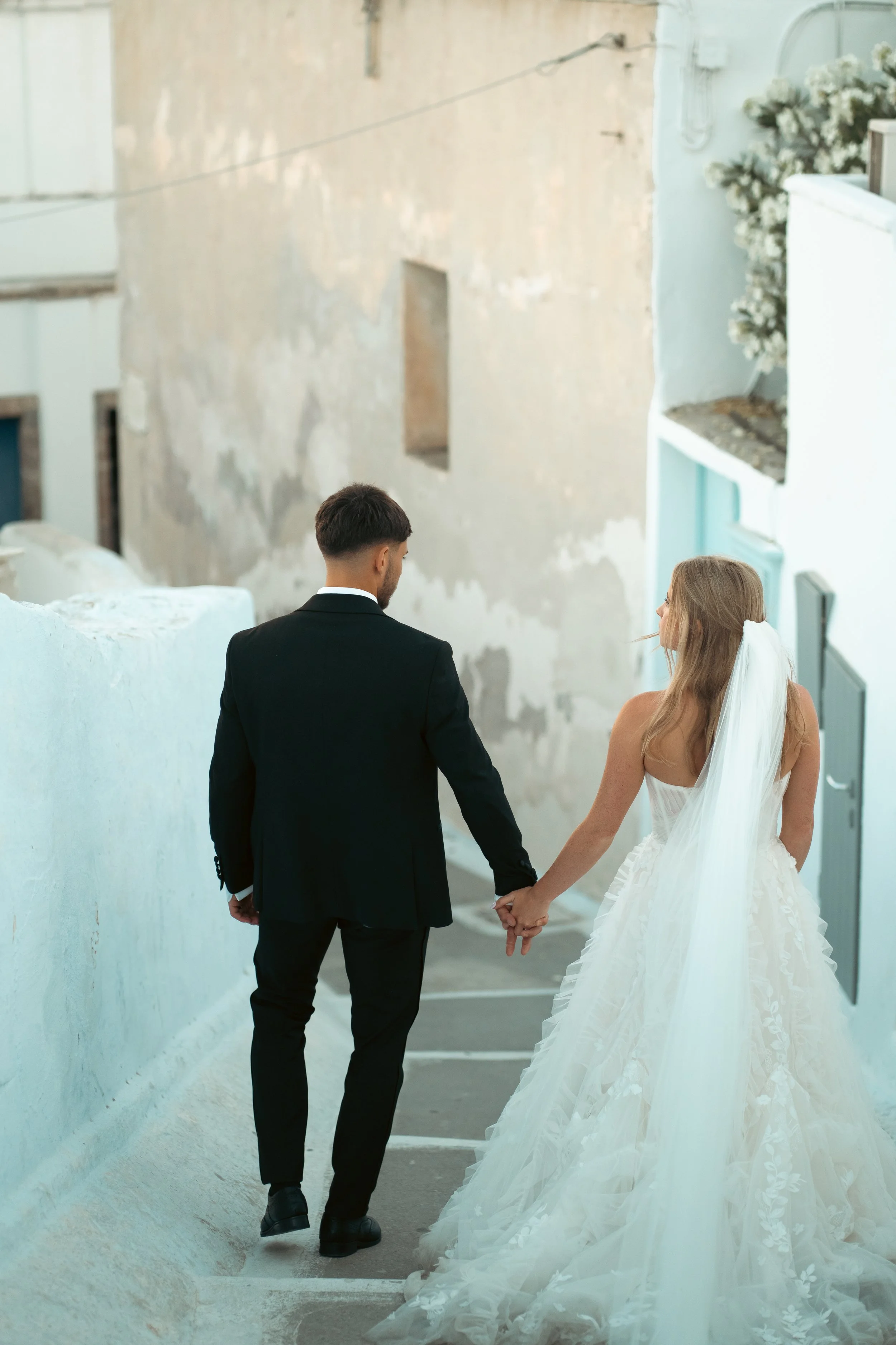 A bride and groom holding hands while walking down a narrow street with white walls, the bride in a strapless wedding gown with a long train and veil, and the groom in a black suit.