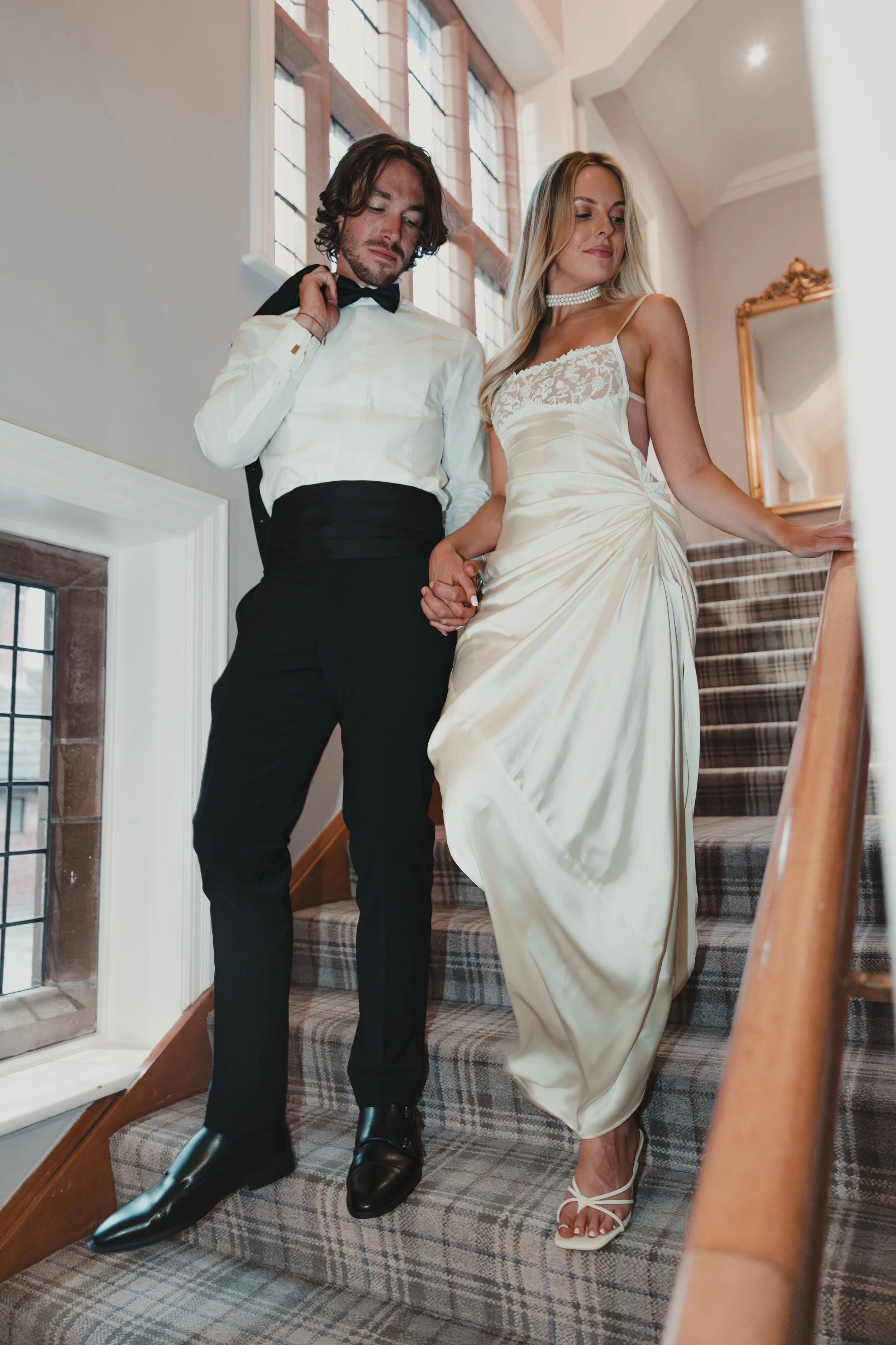 A man and woman in formal attire, holding hands, descending a staircase in a well-lit indoor setting, possibly at a wedding.