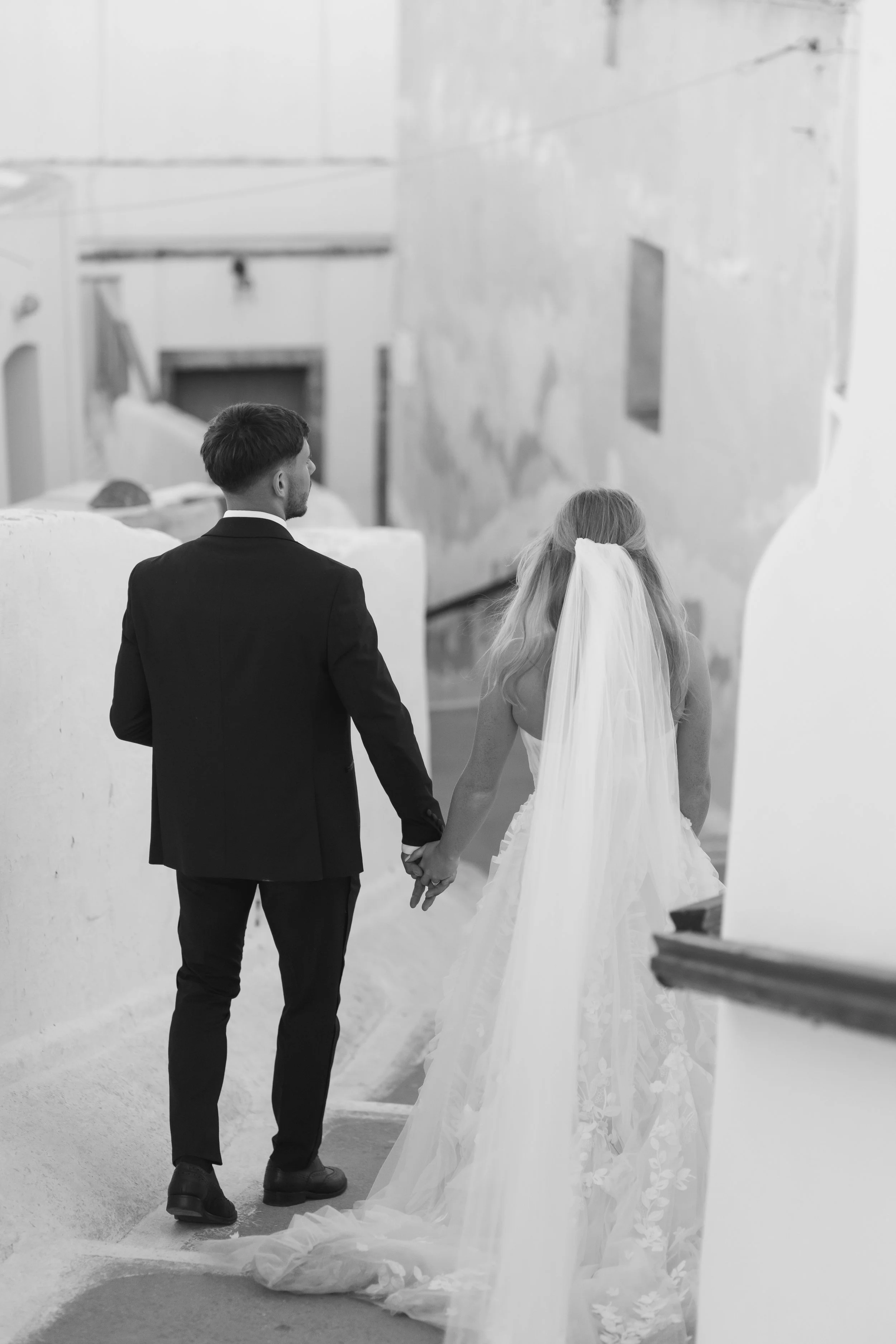 A bride and groom holding hands, walking down a staircase in a white-washed building, with the bride wearing a long veil and gown, and the groom in a suit, in a black-and-white photo.