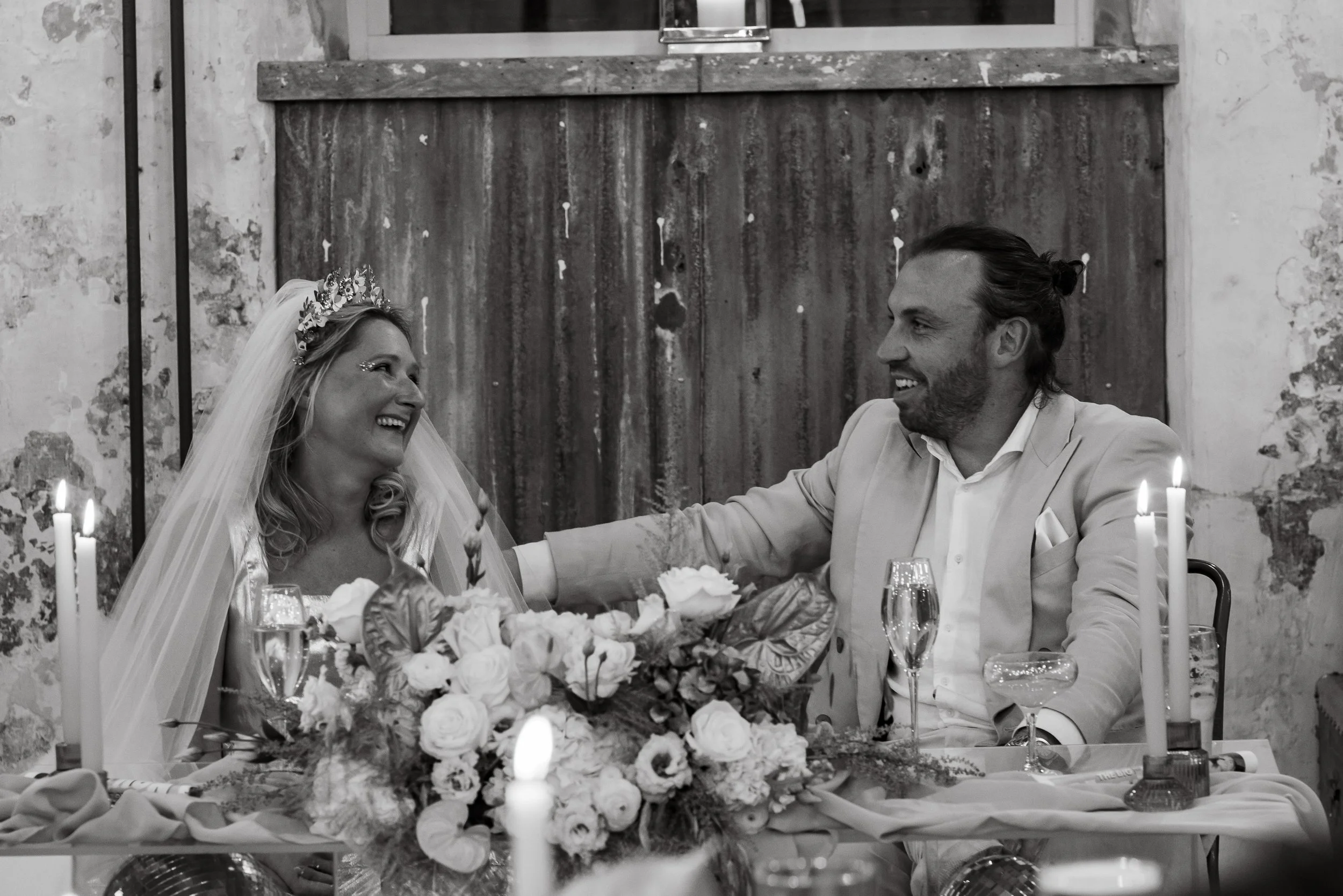 A bride and groom smiling at each other during a wedding reception, seated at a table with flowers, candles, and champagne glasses at Holmes Mill