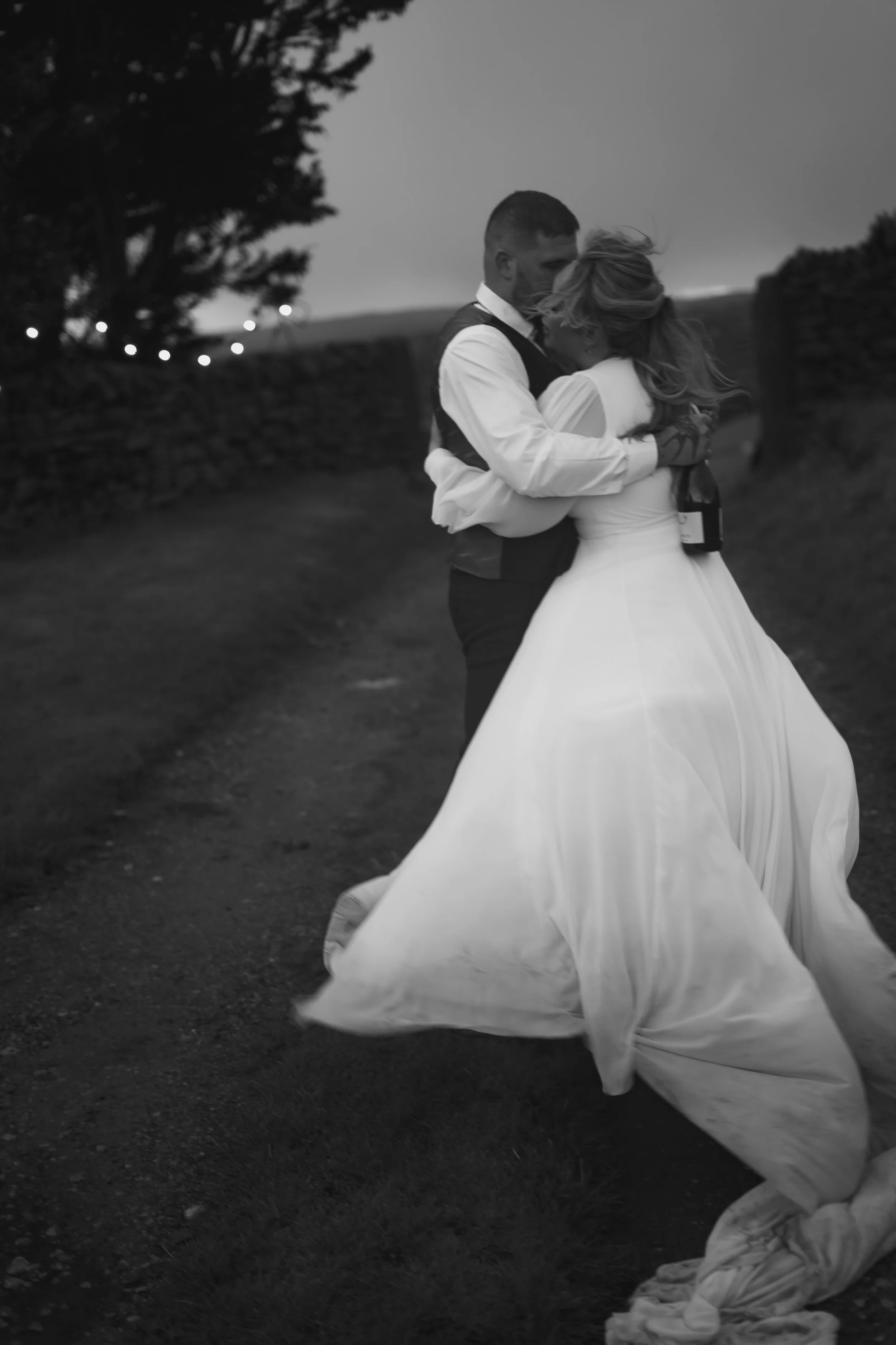 A couple in wedding attire embracing outdoors during dusk, with the bride wearing a flowing white gown and the groom in a waistcoat and shirt, holding a bottle of champagne.