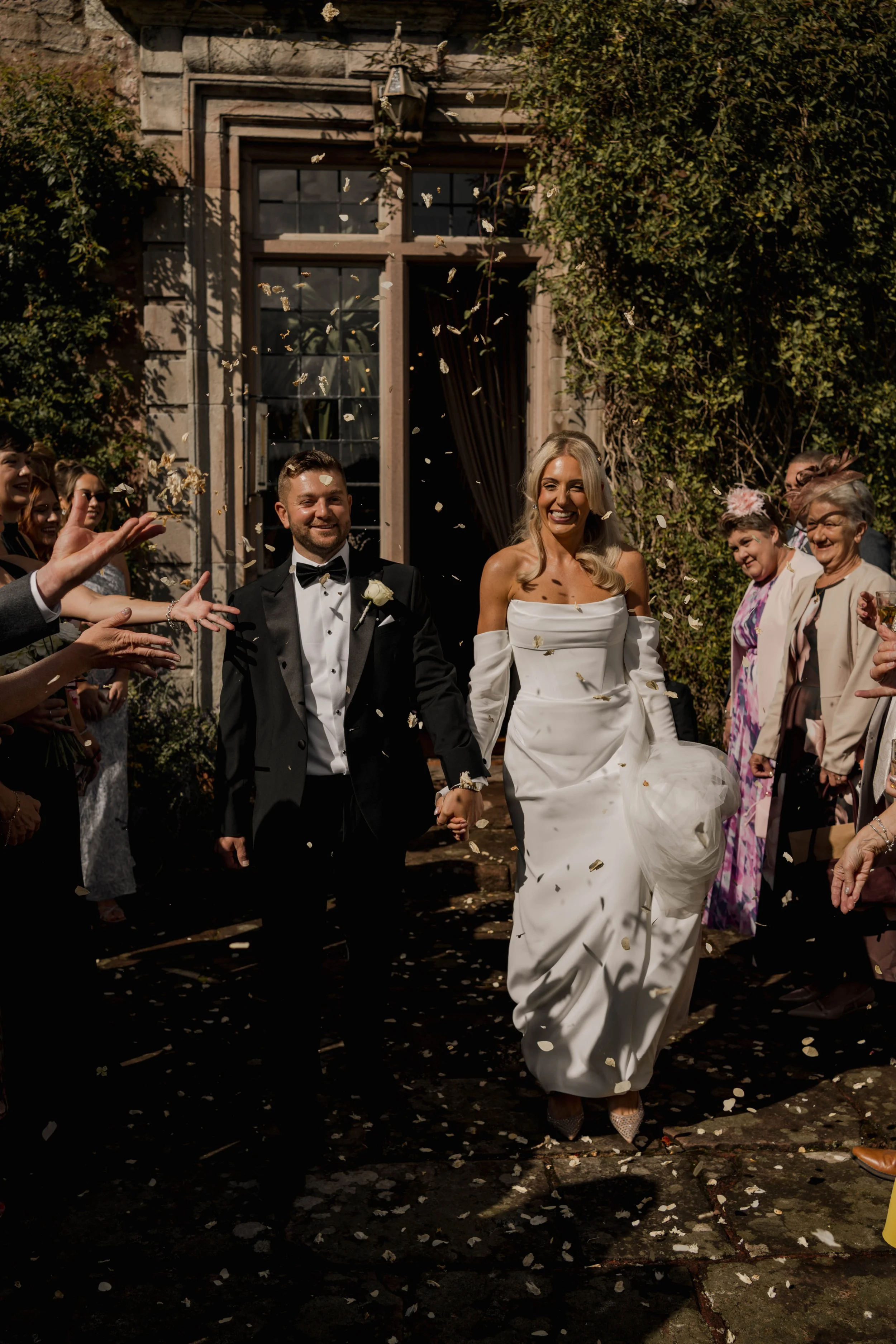 A newlywed couple holding hands and smiling as they walk away from a wedding reception, surrounded by friends and family throwing confetti outside a rustic building.