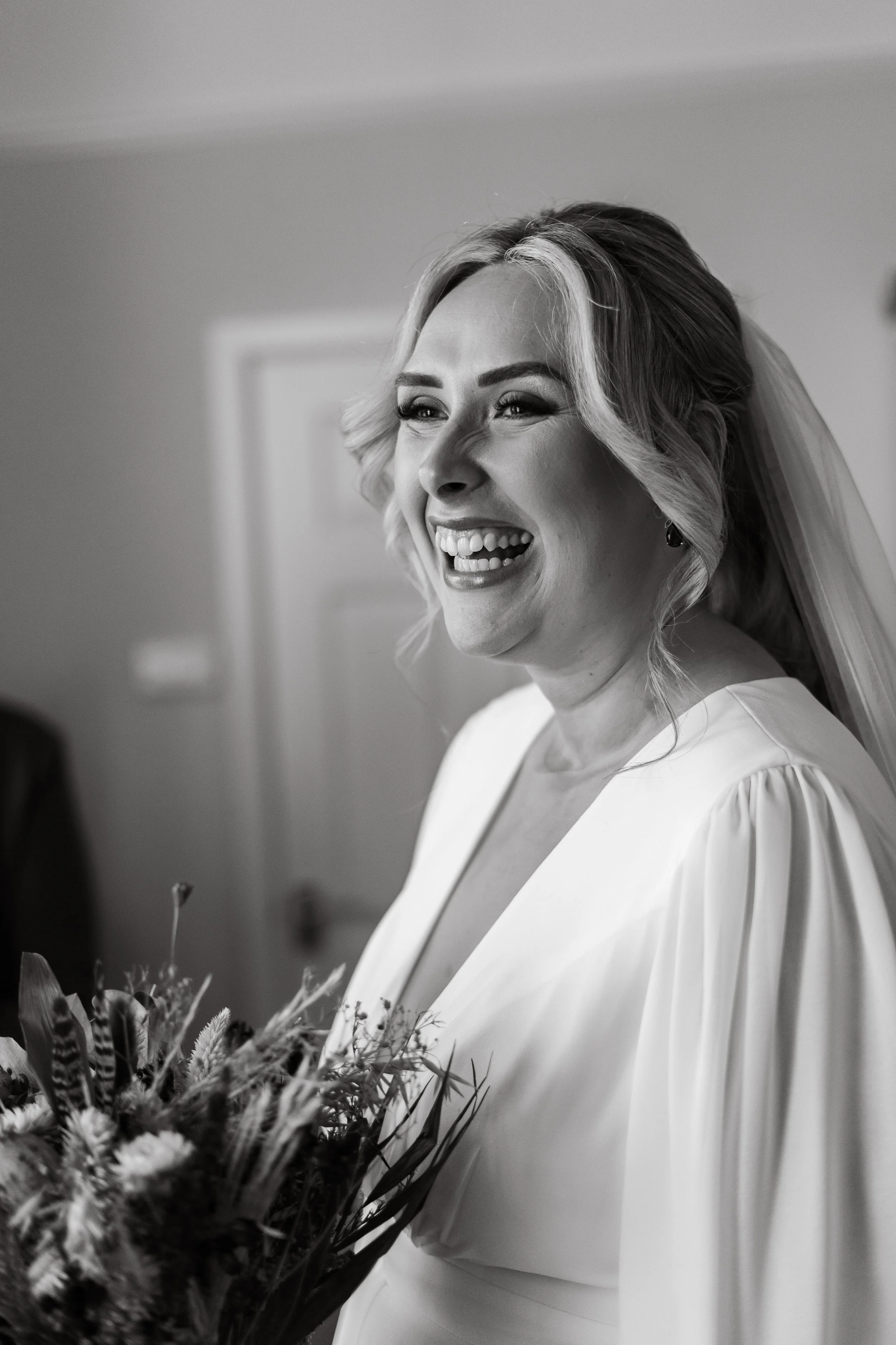 Black and white photo of a smiling bride with blonde hair in a wedding dress, holding a bouquet of flowers.