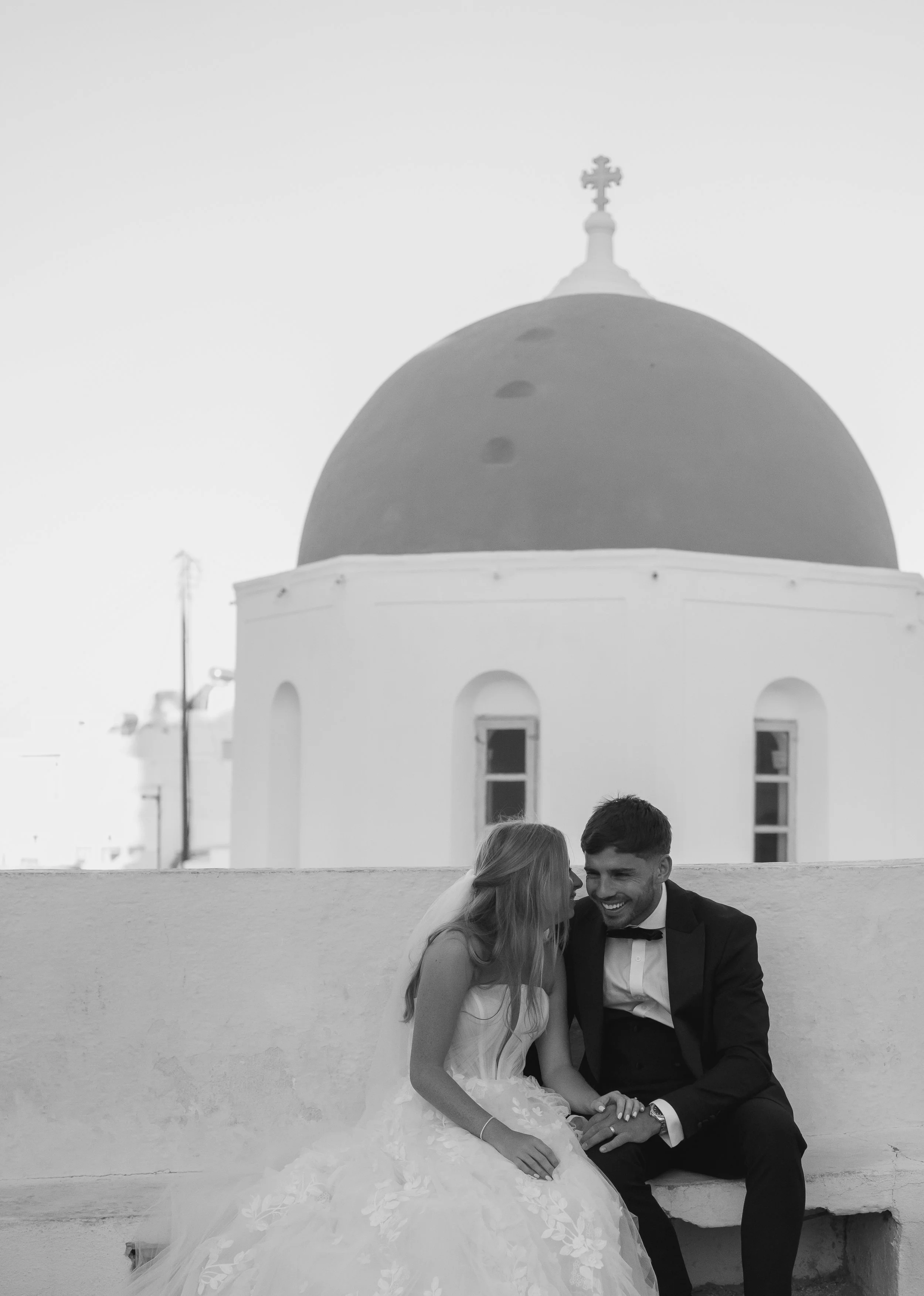 Black and white photo of a bride and groom sitting on a bench, sharing a happy moment, with a white building and a dome with a cross in the background.