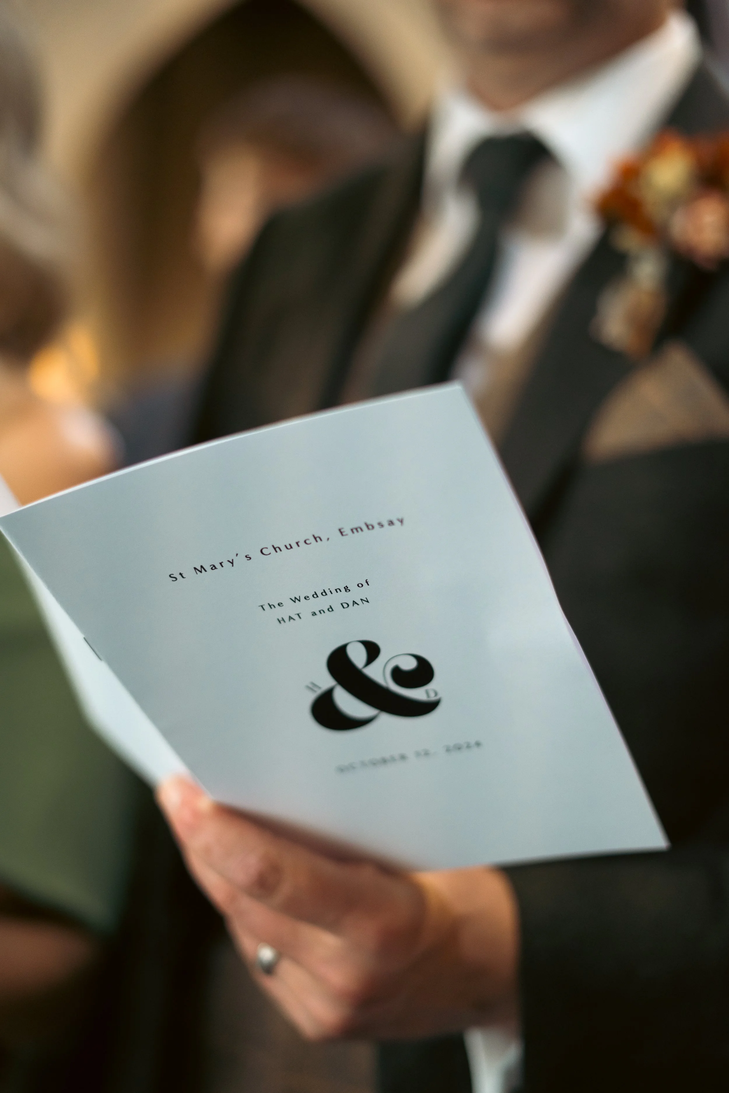 A person in formal attire holding a wedding program for the wedding of Hat and Dan at St. Mary's Church, Embsay, dated October 2, 2022.