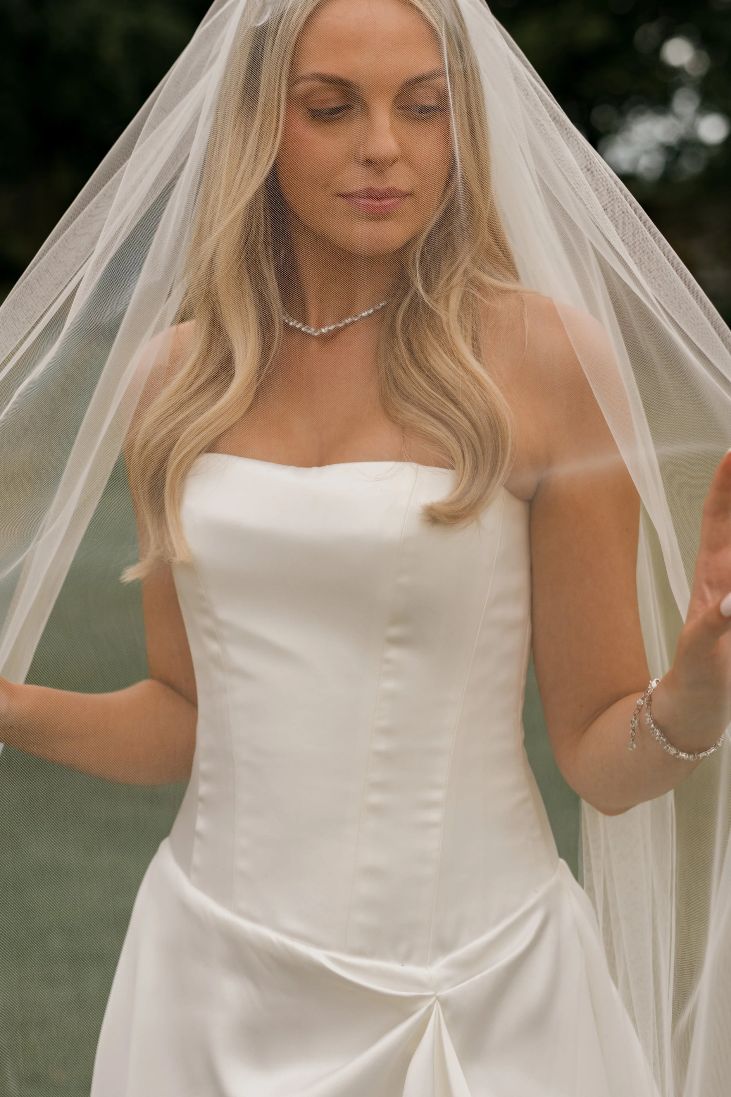 A bride in a white strapless wedding dress, wearing a veil, necklace, and bracelet, standing outdoors with a dark green background.