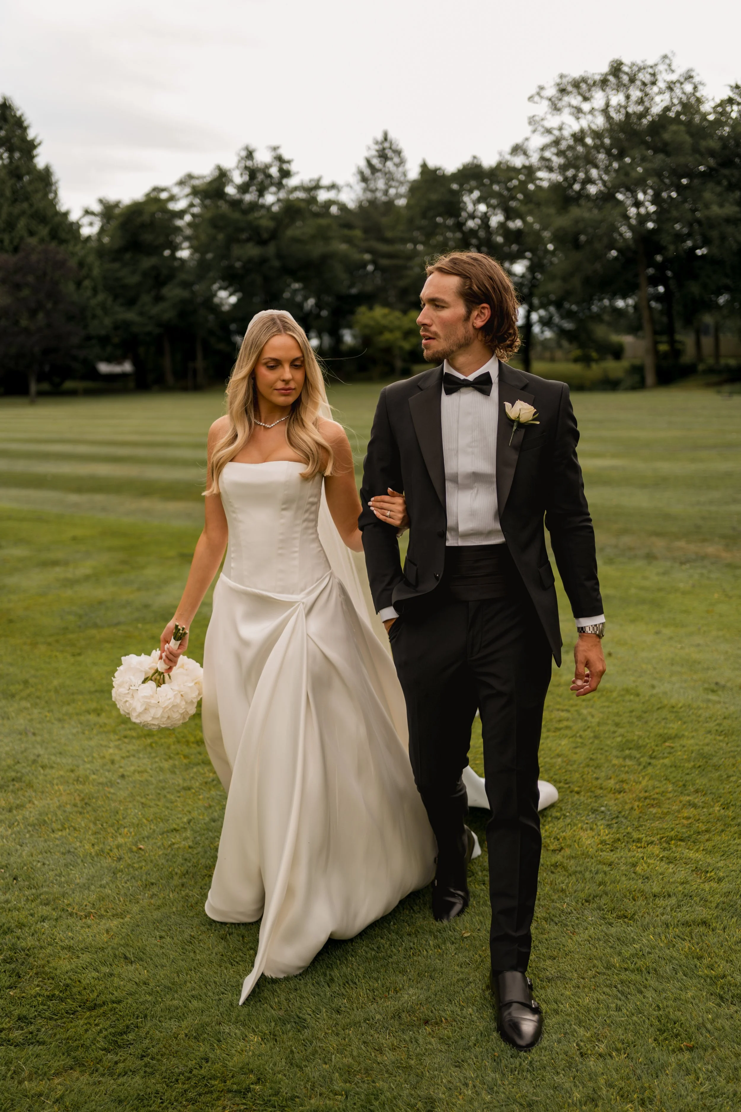 A bride and groom walking arm in arm on a grassy field during their wedding, with trees in the background.