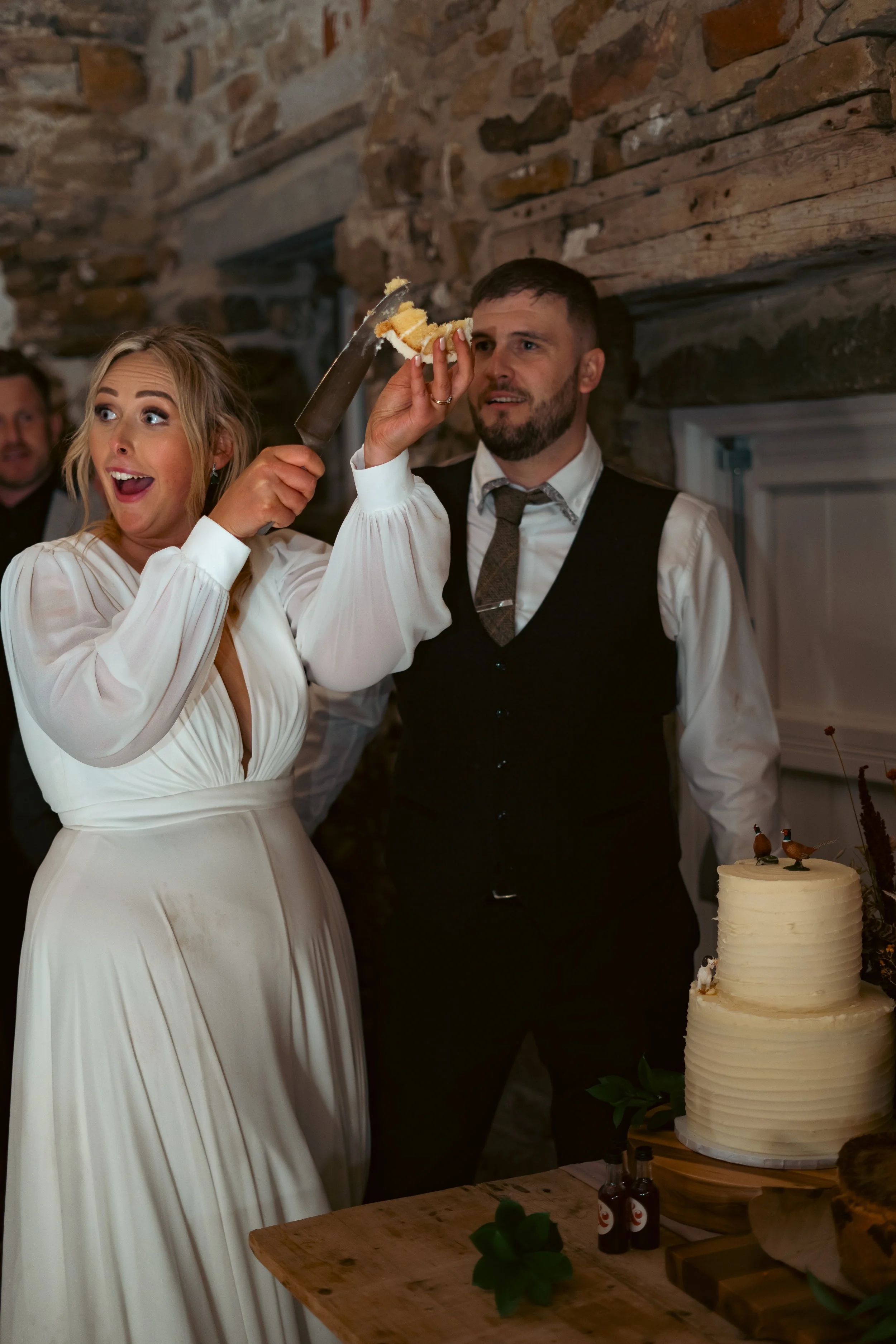 Bride and groom at wedding cake cutting ceremony, woman smiling as cake is being cut with a sword, groom holding cake, rustic indoor setting.