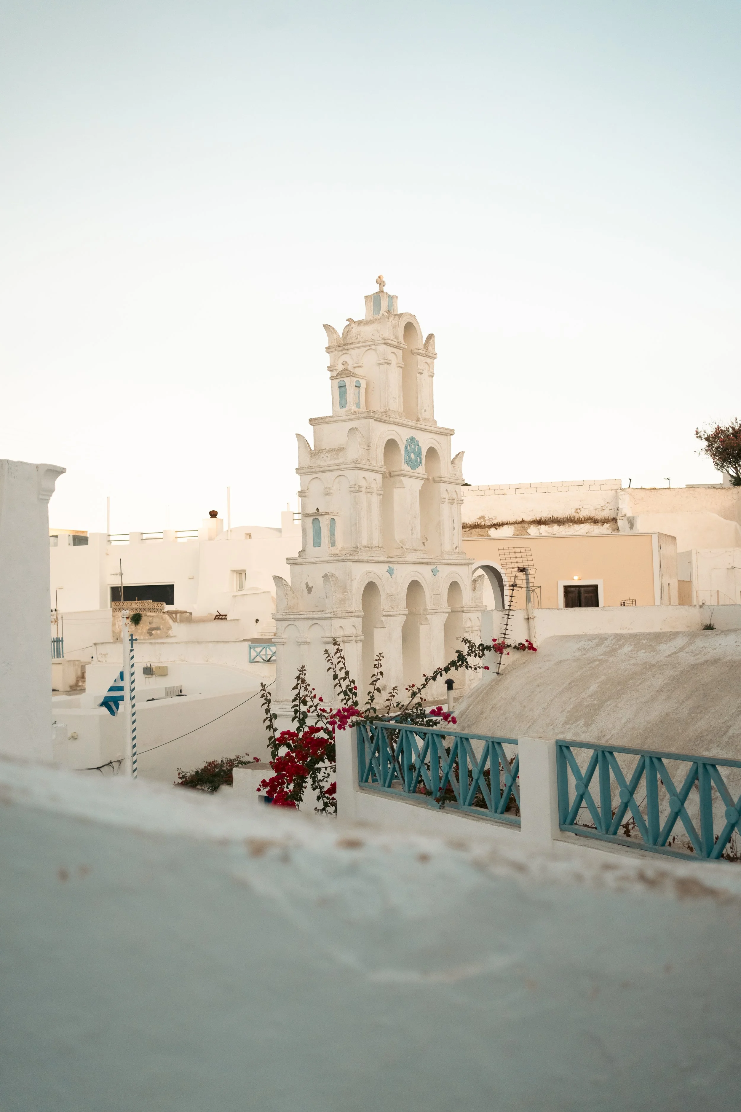 A white tower with blue accents in a Mediterranean village, surrounded by white buildings, pink flowers, and blue railings, under a clear sky.
