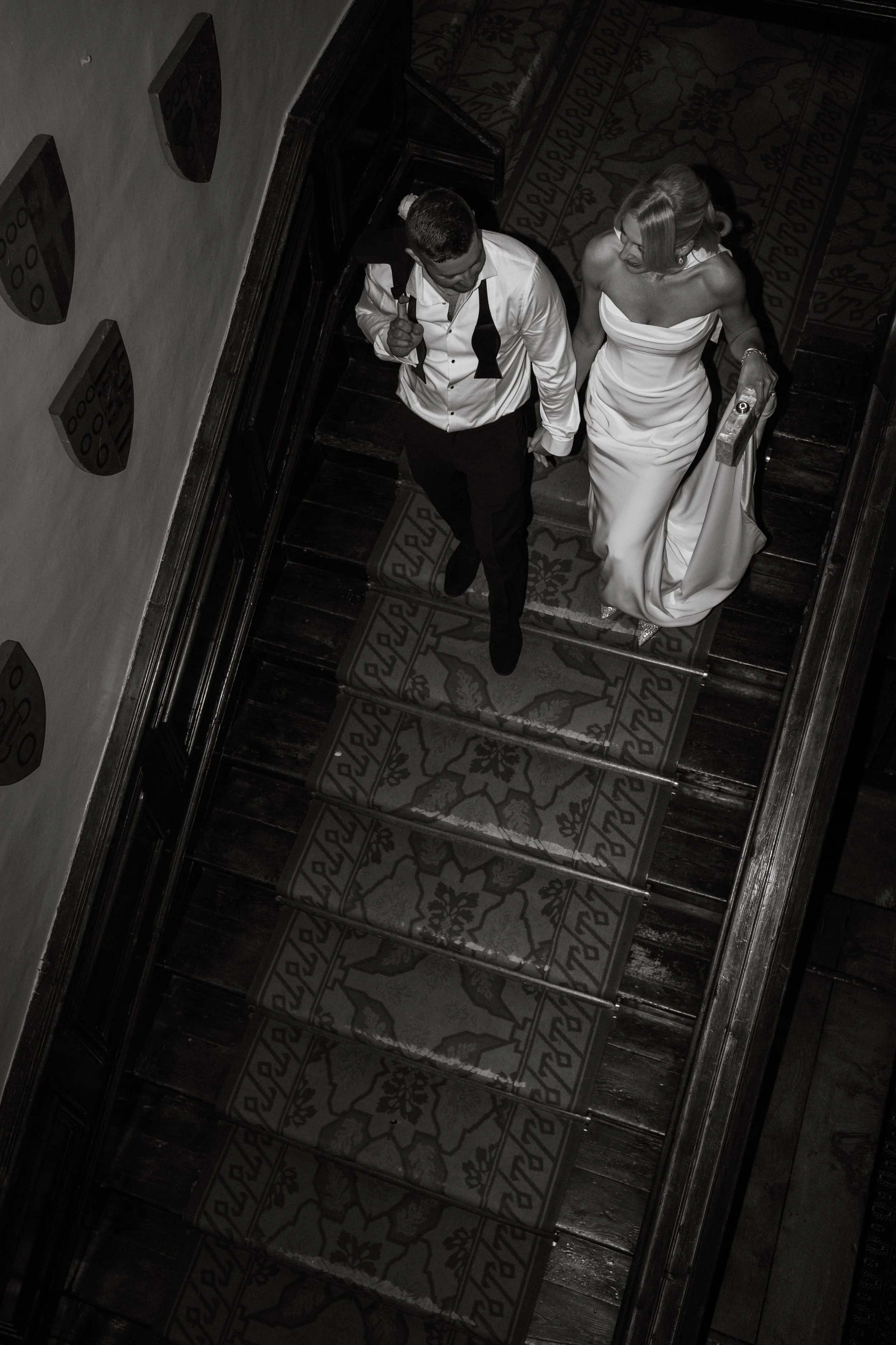 A bride and groom walking up a staircase, holding hands. The groom is carrying a jacket over his shoulder and wearing a white shirt and black pants. The bride is in a strapless wedding gown, holding a bouquet, with her hair styled up.