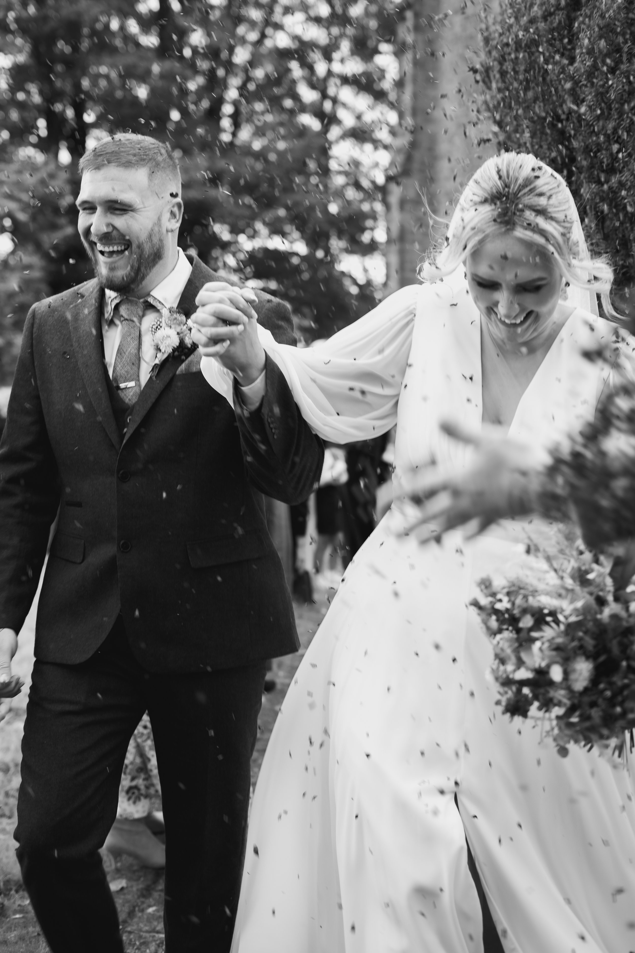 Black and white photo of a newlywed couple, smiling and holding hands, celebrating outdoors with confetti flying around them.