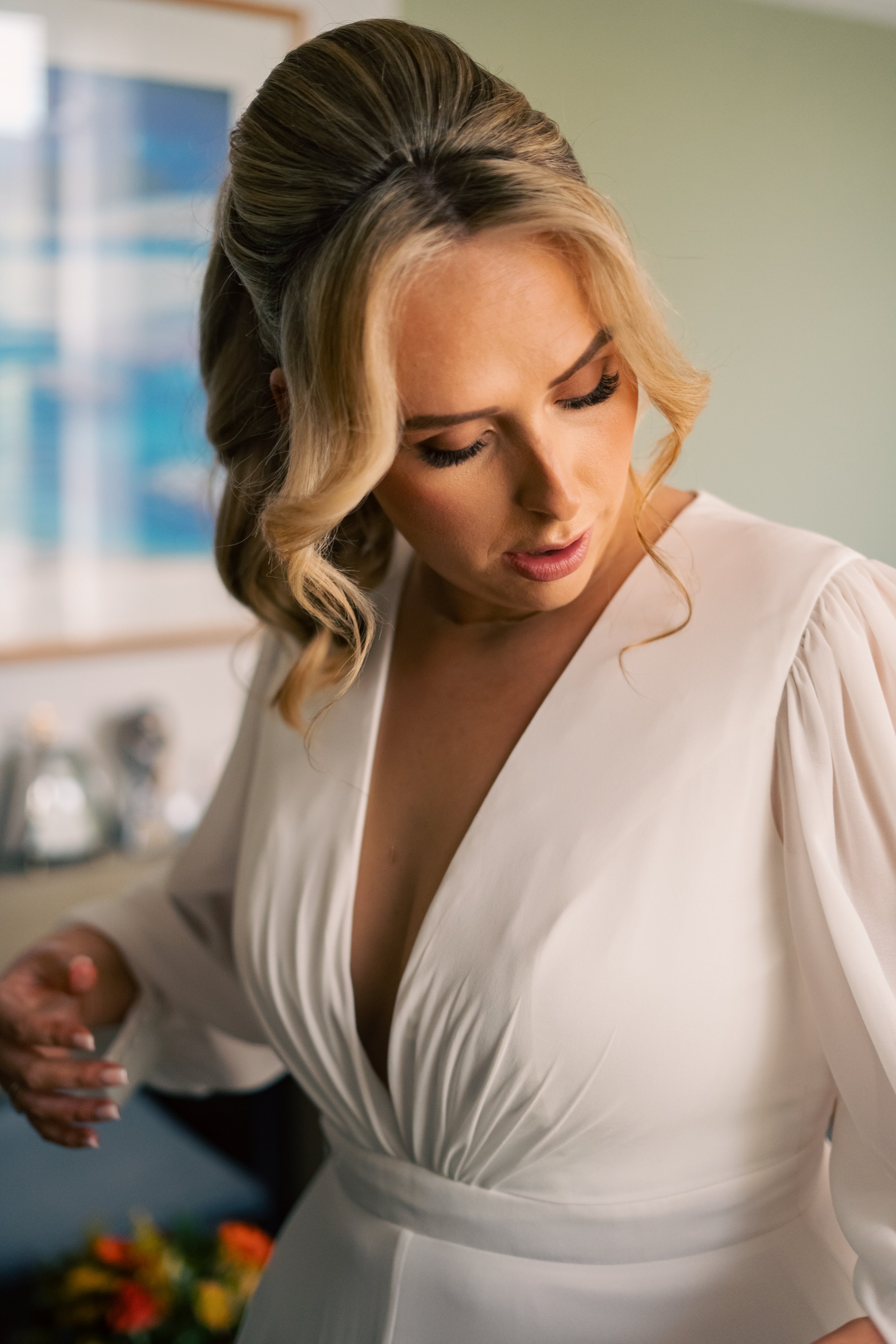 A bride in a white dress looking down, with styled blonde hair and makeup, in an indoor setting.