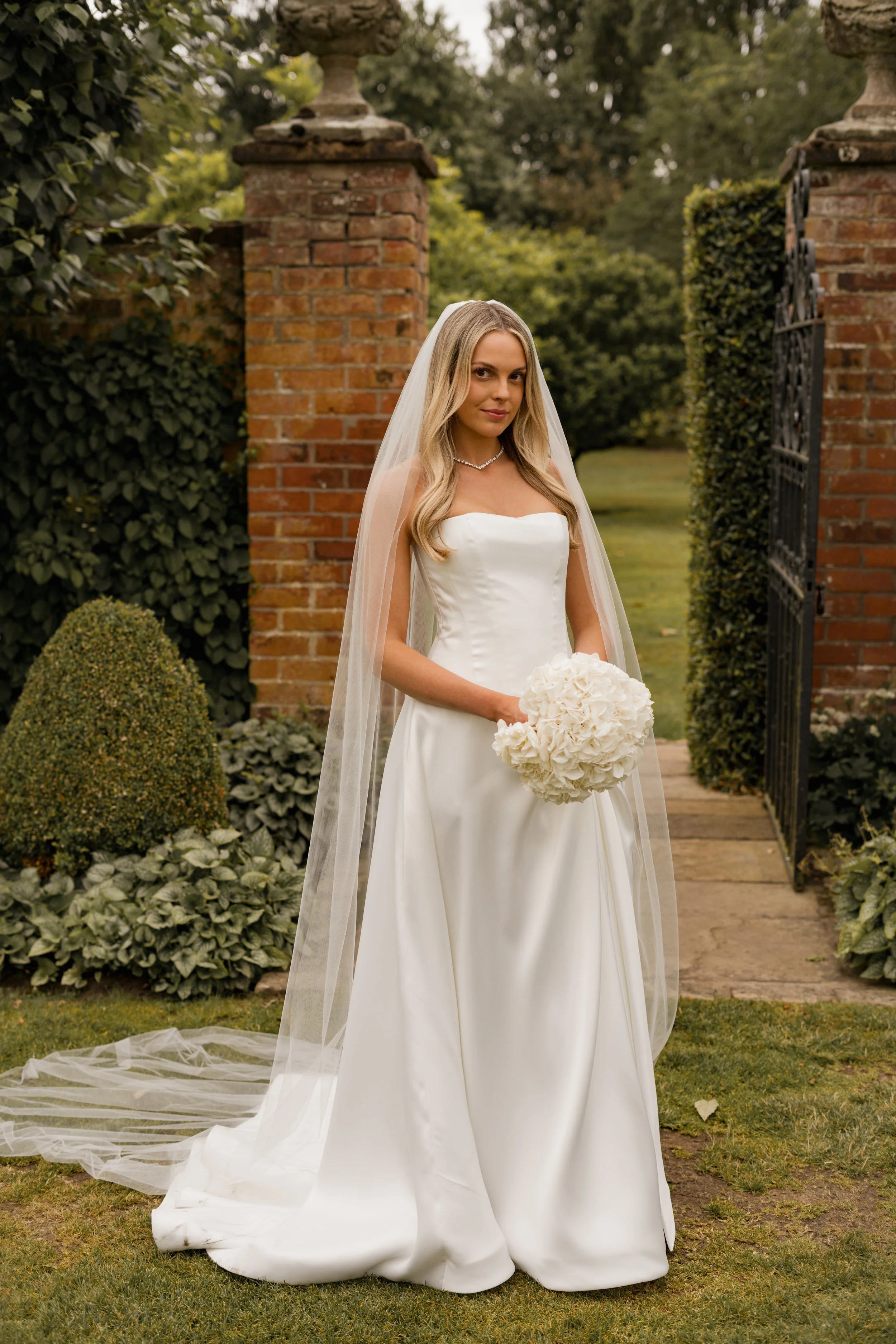 A bride in a white wedding gown holding a bouquet of white flowers, standing outdoors near a brick gate with greenery in the background.