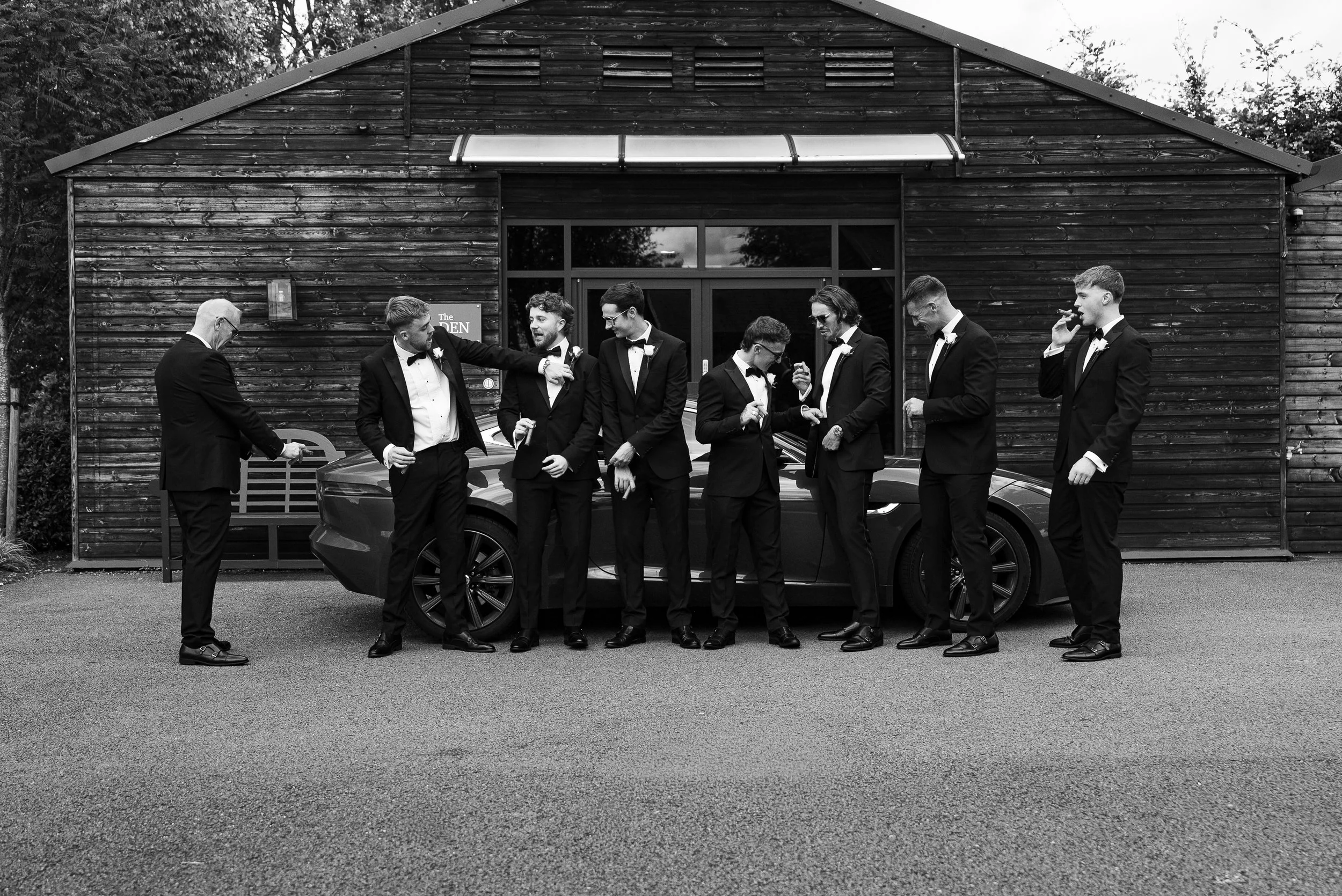 A group of nine men dressed in tuxedos standing in front of a car and a wooden building, with some talking and one on the phone, in black and white.