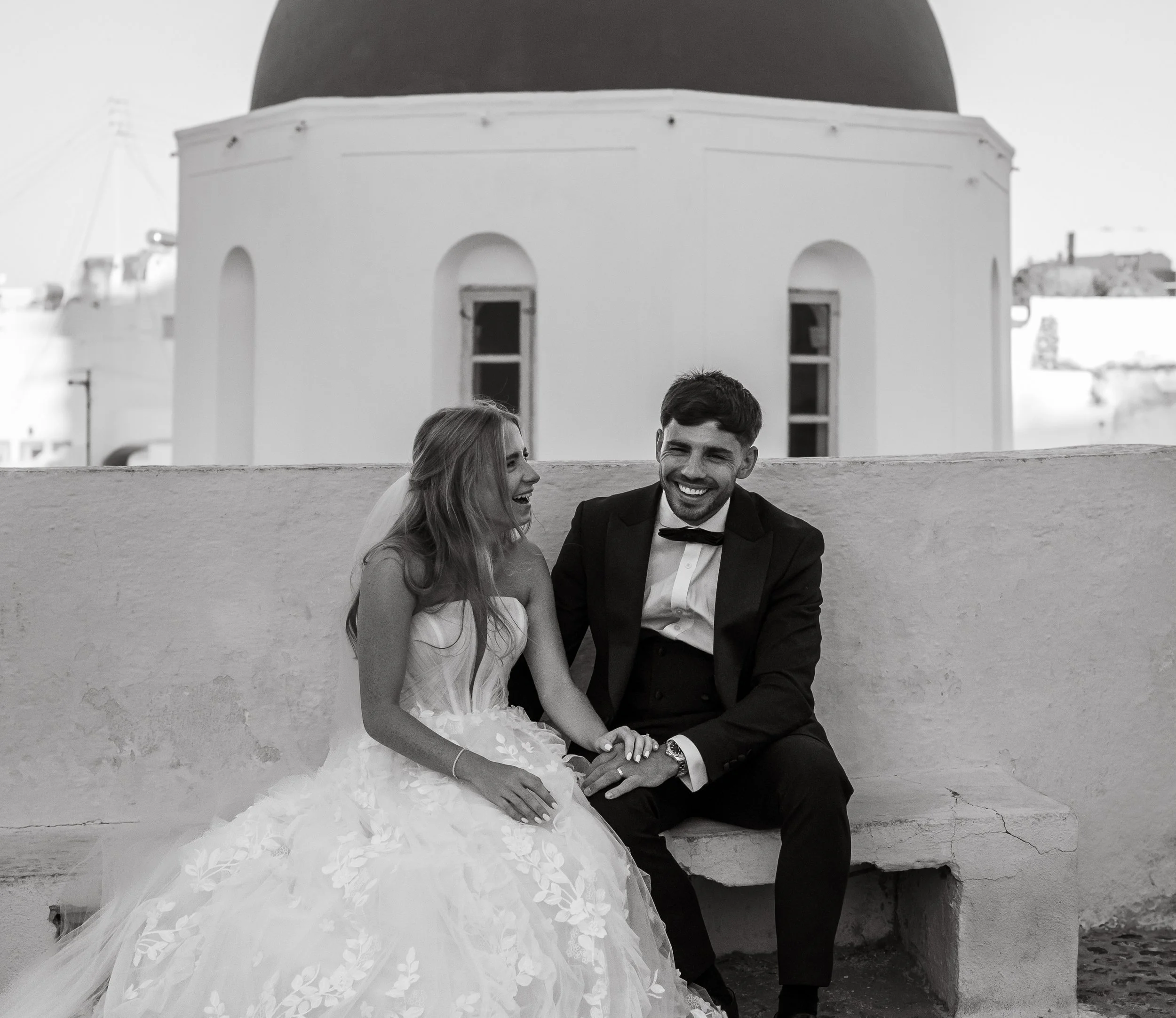 A happy bride and groom sitting on a bench, laughing and holding hands, with a white building featuring arched windows and a dome in the background.