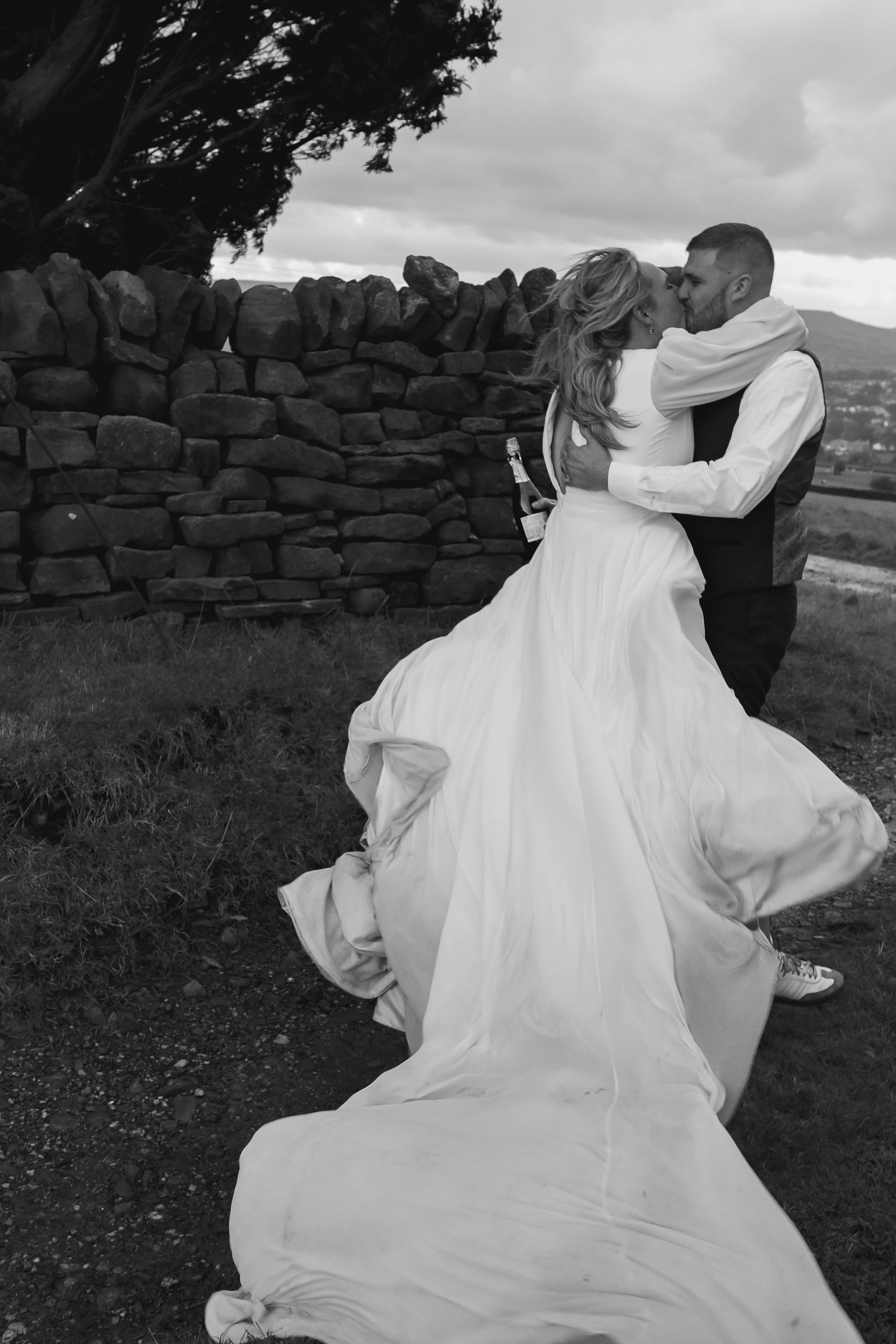 A black and white photo of a couple in wedding attire hugging and kissing outdoors near a stone wall, with a cloudy sky and distant hills in the background.