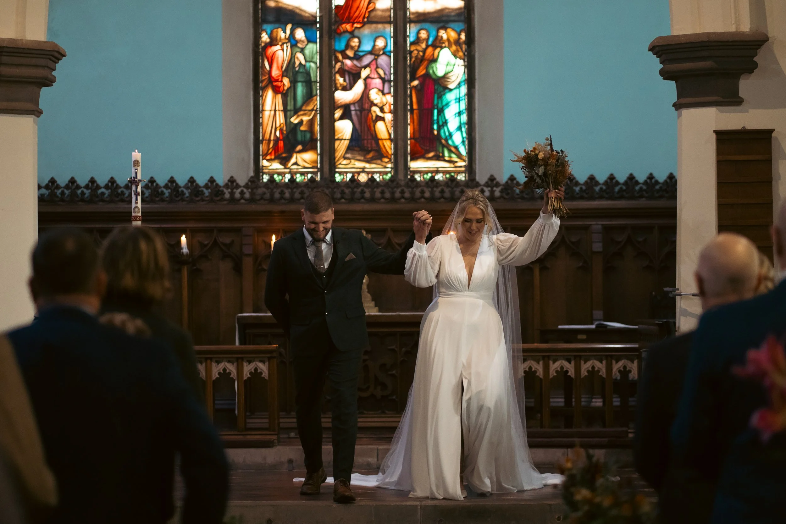 A bride and groom holding hands and walking down the aisle in a church after their wedding, with stained glass windows in the background and guests seated in front.