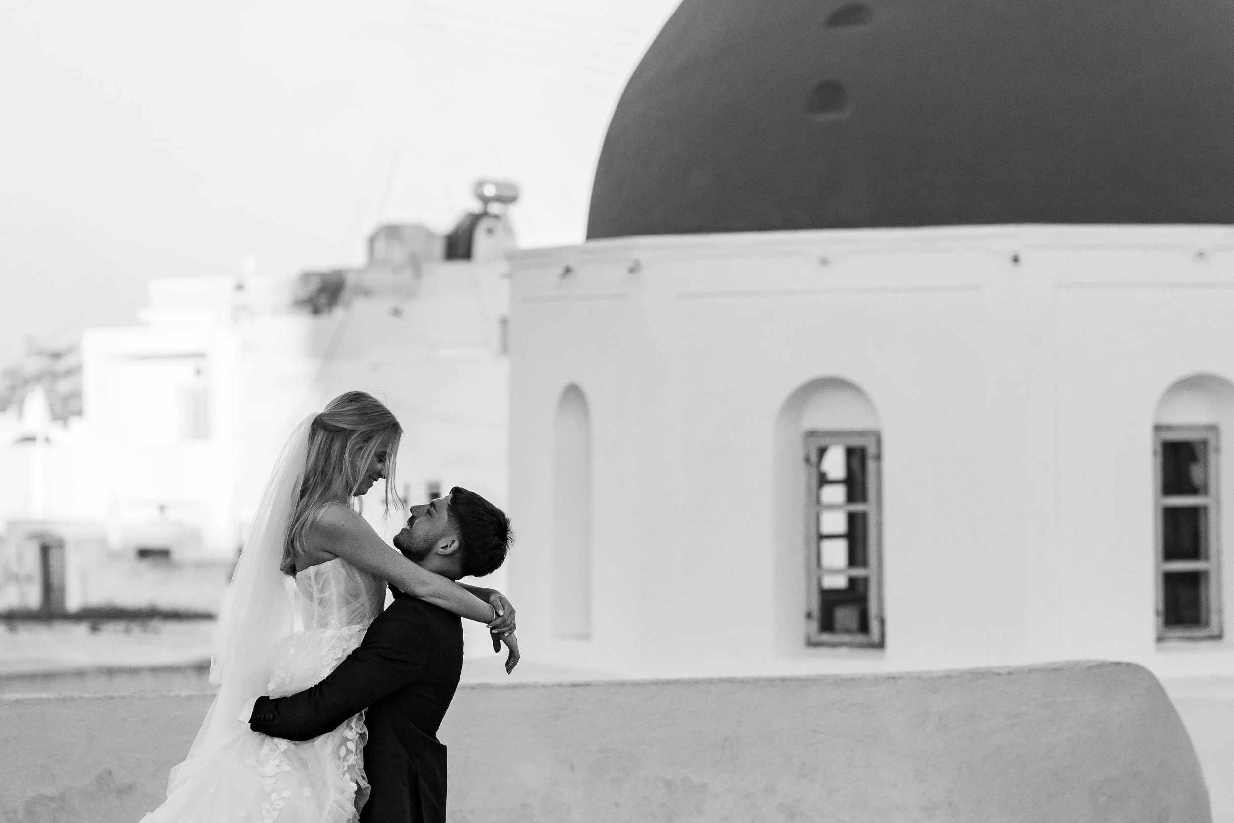 A black-and-white photo of a wedding couple with the man lifting the woman in his arms, standing outdoors near white buildings with arched windows and a domed roof in the background.