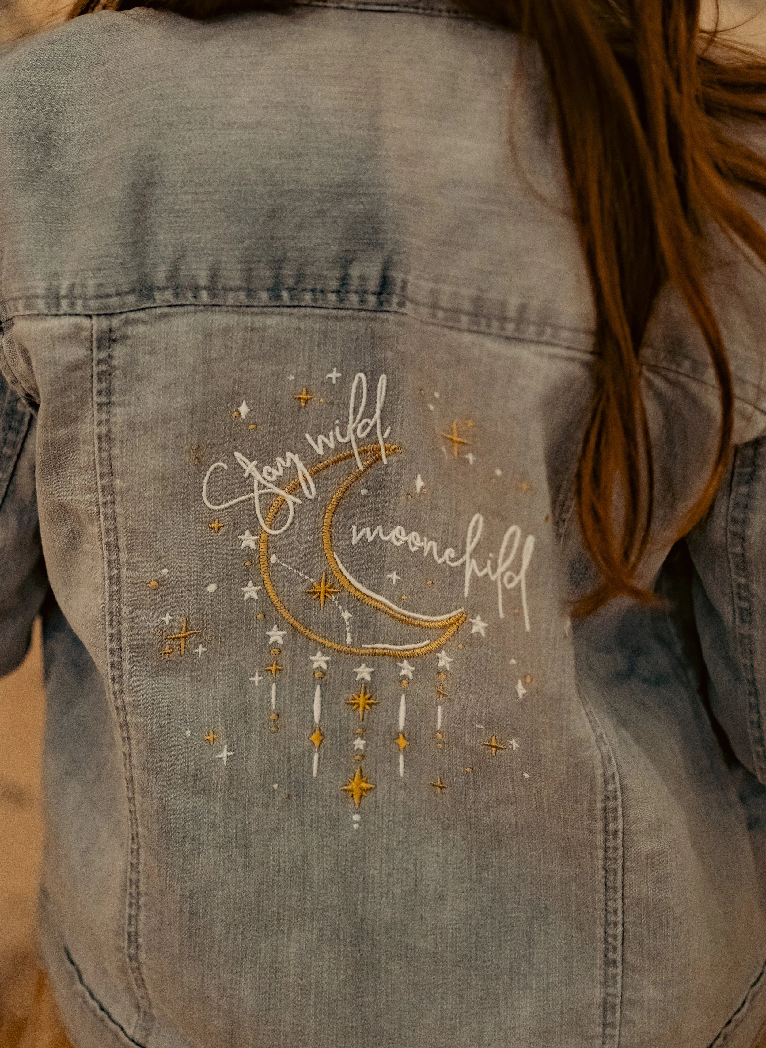 Back of a gray denim jacket with embroidered crescent moon, stars, and hanging tassels, and the words 'Stay wild, moon child'.