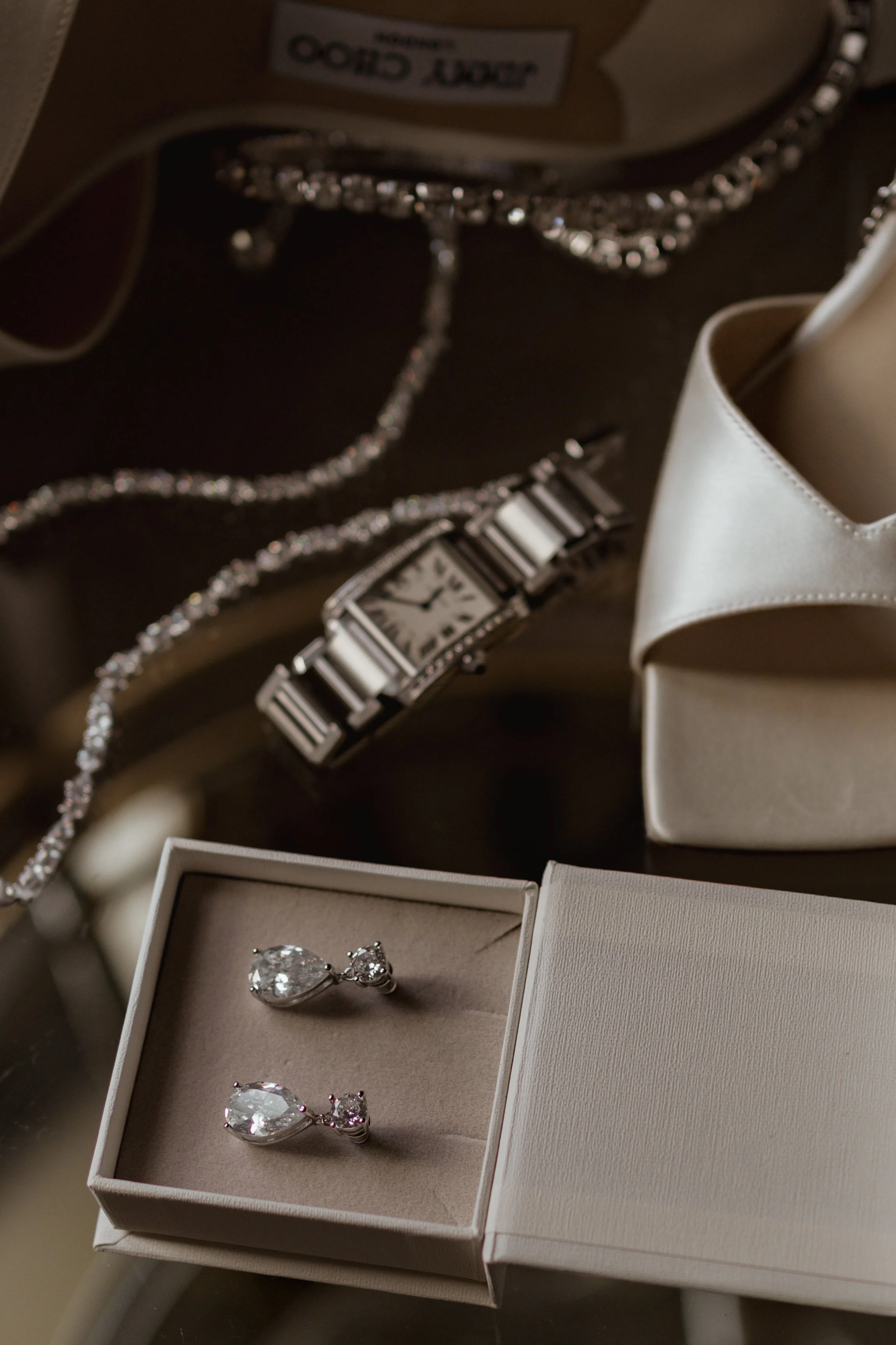 Jewelry including diamond earrings in an open jewelry box, a wristwatch, a silver necklace, and a pair of white shoes on a dark surface.