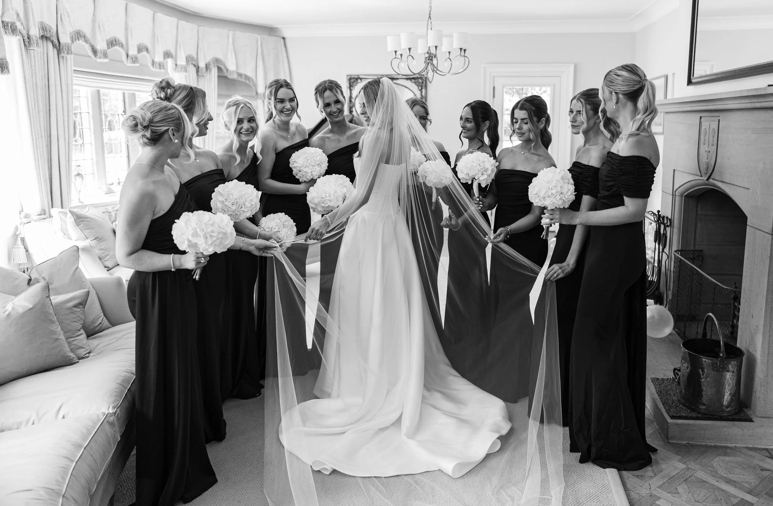 Bride in a wedding gown with veil surrounded by bridesmaids in black dresses holding bouquets in a bright living room.