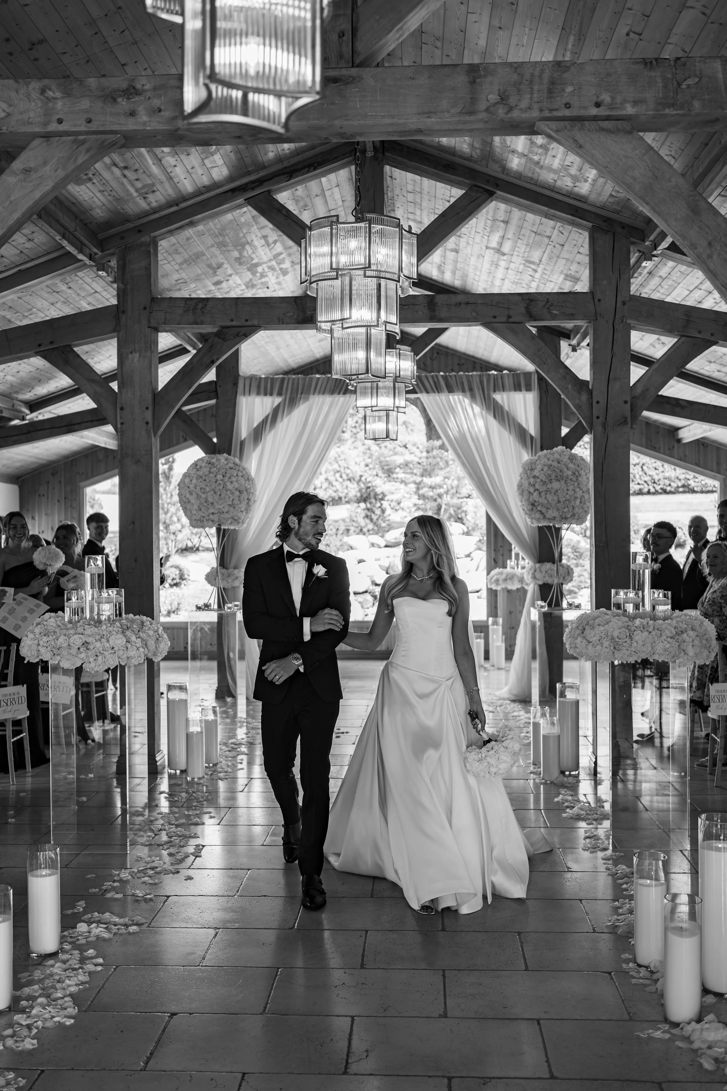 A black and white photo of a bride and groom walking down the aisle inside a rustic wooden wedding venue with chandeliers, floral decorations, and guests sitting on either side.