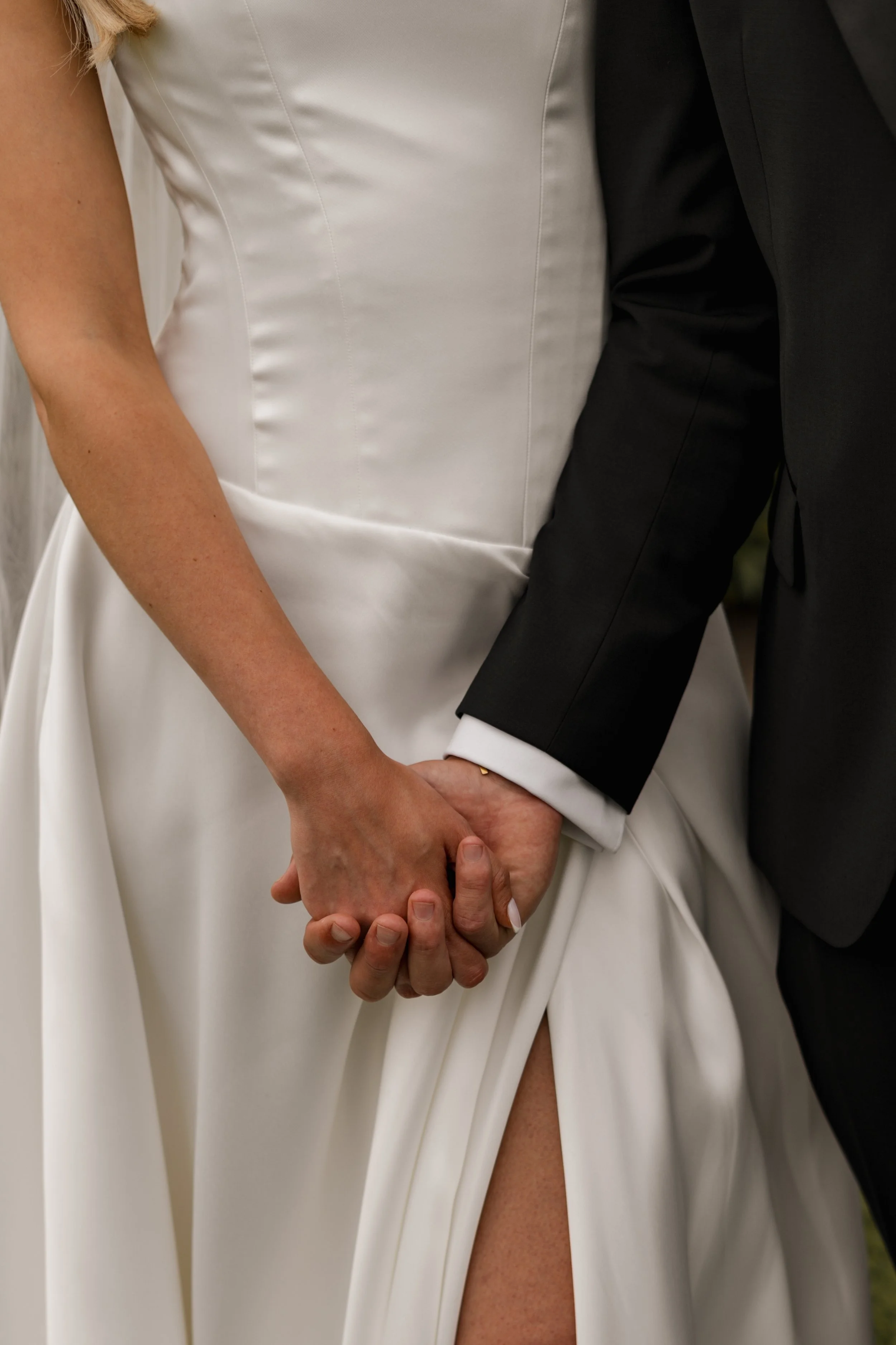 Close-up of a bride and groom holding hands, with the bride wearing a white wedding dress and the groom in a black suit.