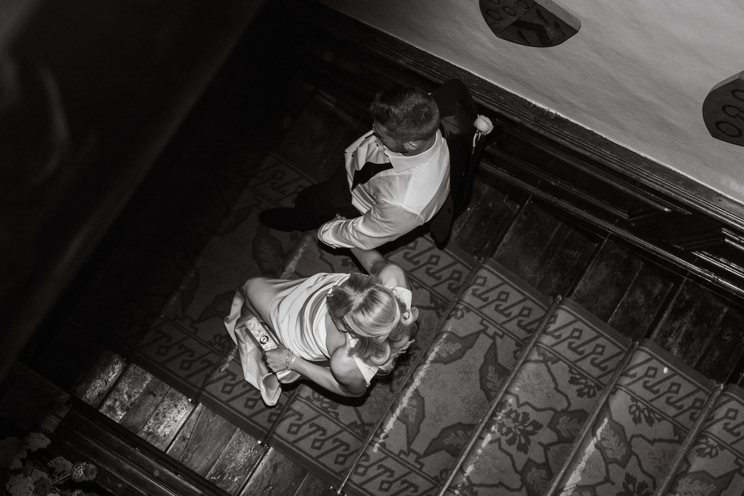 A black and white photo of a man and woman sitting on a staircase, viewed from above. The man is wearing a white shirt with black accents, sitting at the top of the stairs, looking down. The woman, with blonde hair, is sitting on a patterned carpeted section of the stairs, holding a drink and looking to the side.