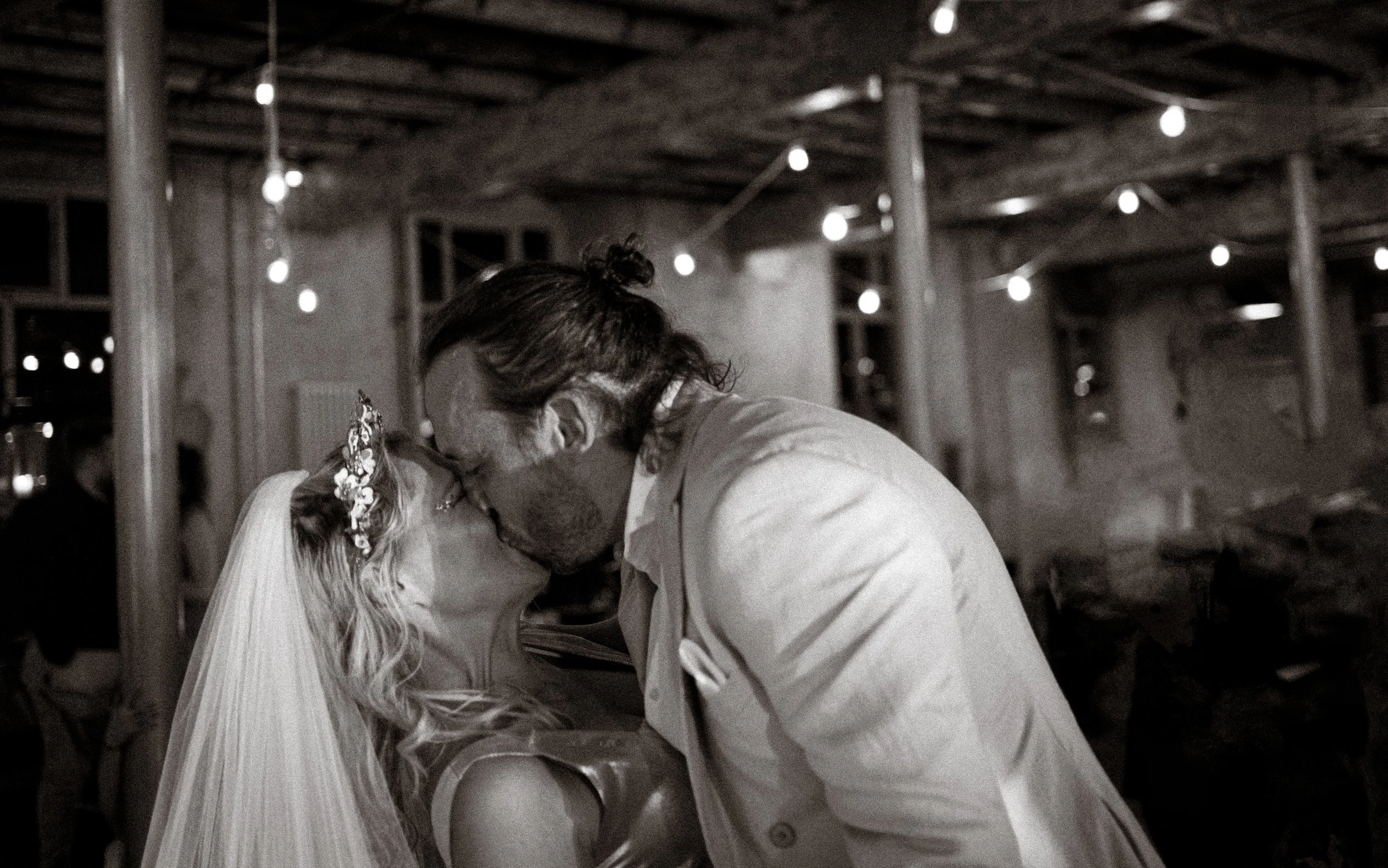 A black and white photo of a couple kissing at their wedding, with the bride wearing a veil and floral headpiece, and the groom wearing a light jacket, in a rustic setting with hanging string lights and wooden beams.