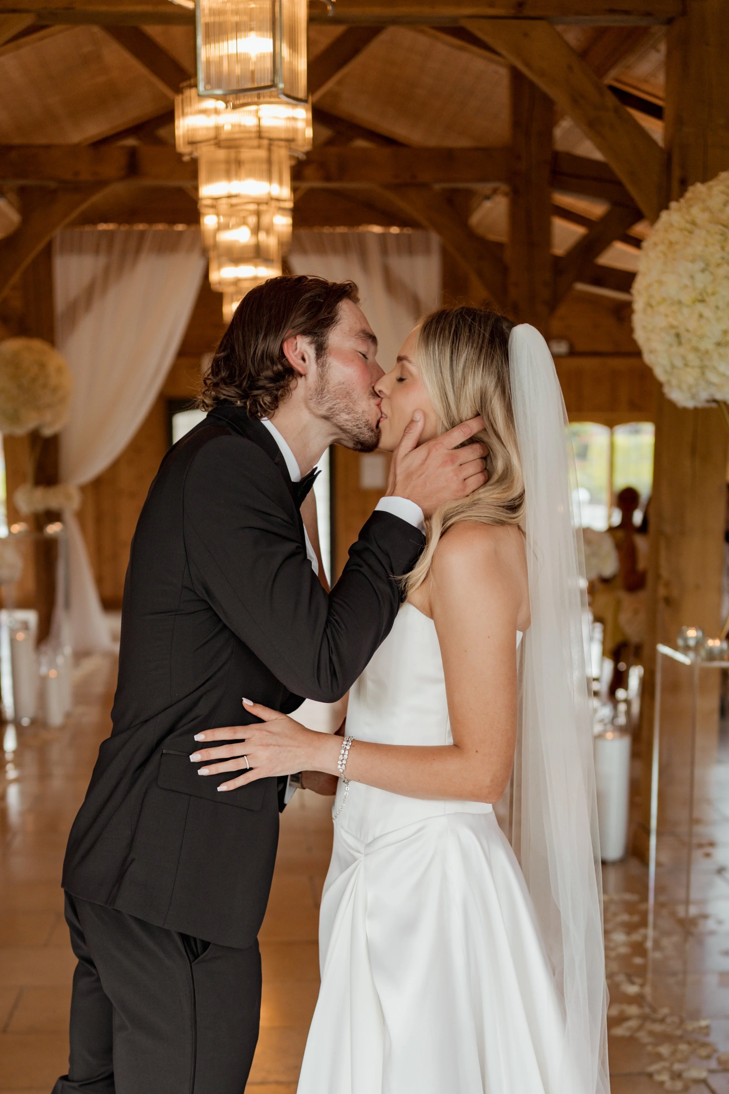 A newlywed couple shares a kiss in a rustic wooden venue decorated with white floral arrangements and draped curtains.