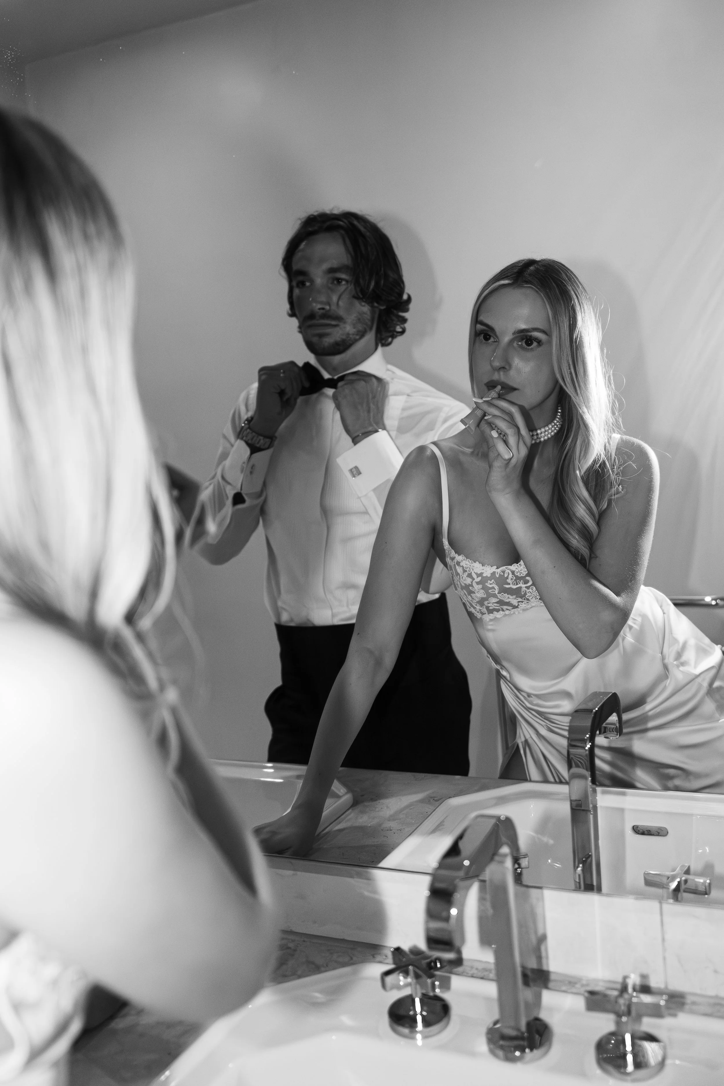 A woman in a silk slip dress and pearl necklace is applying lipstick in front of a mirror, while a man in a white shirt and bow tie adjusts his bow tie in the background.