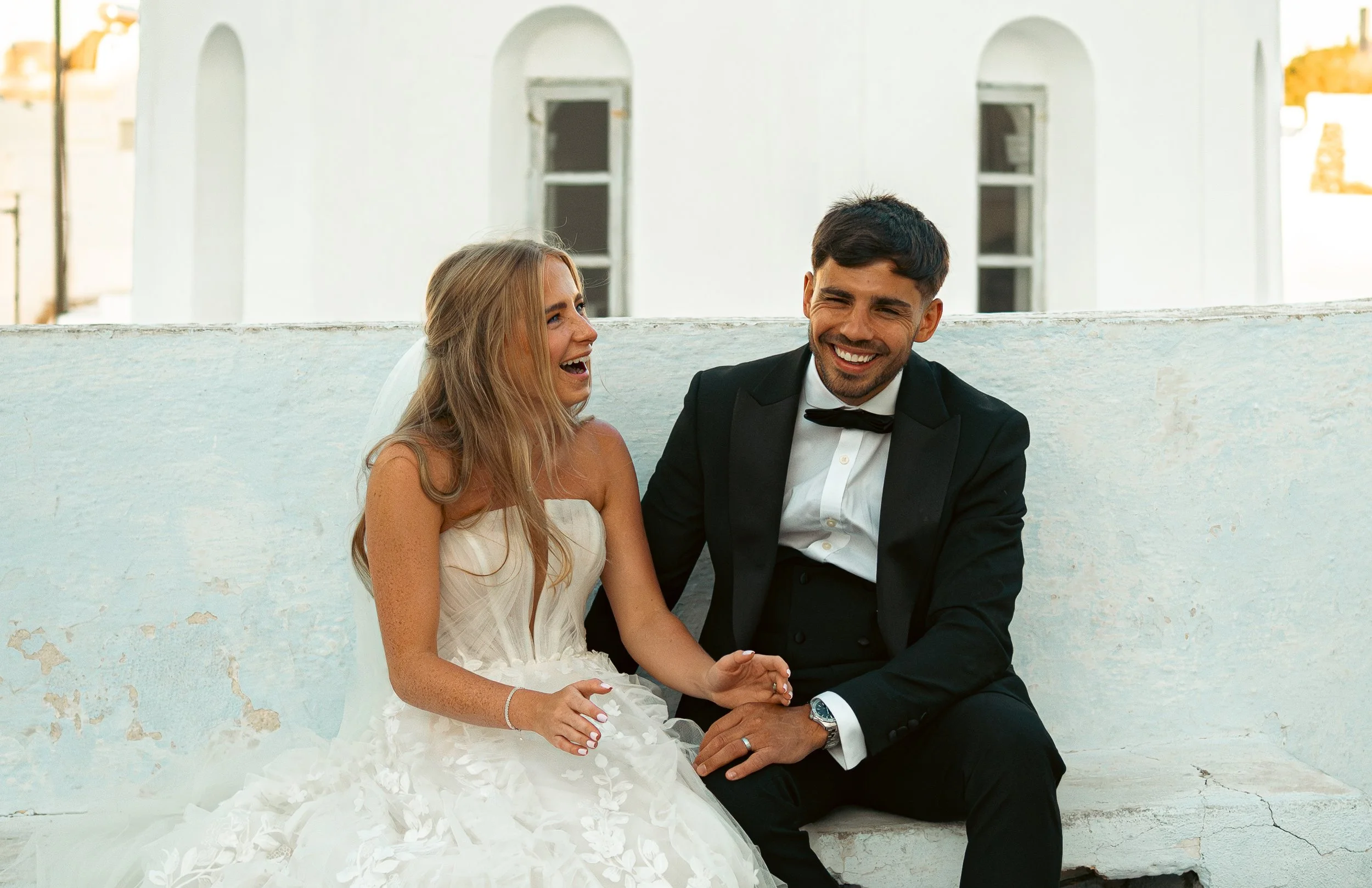 Bride and groom sitting on a white bench, laughing and enjoying their wedding day outdoors